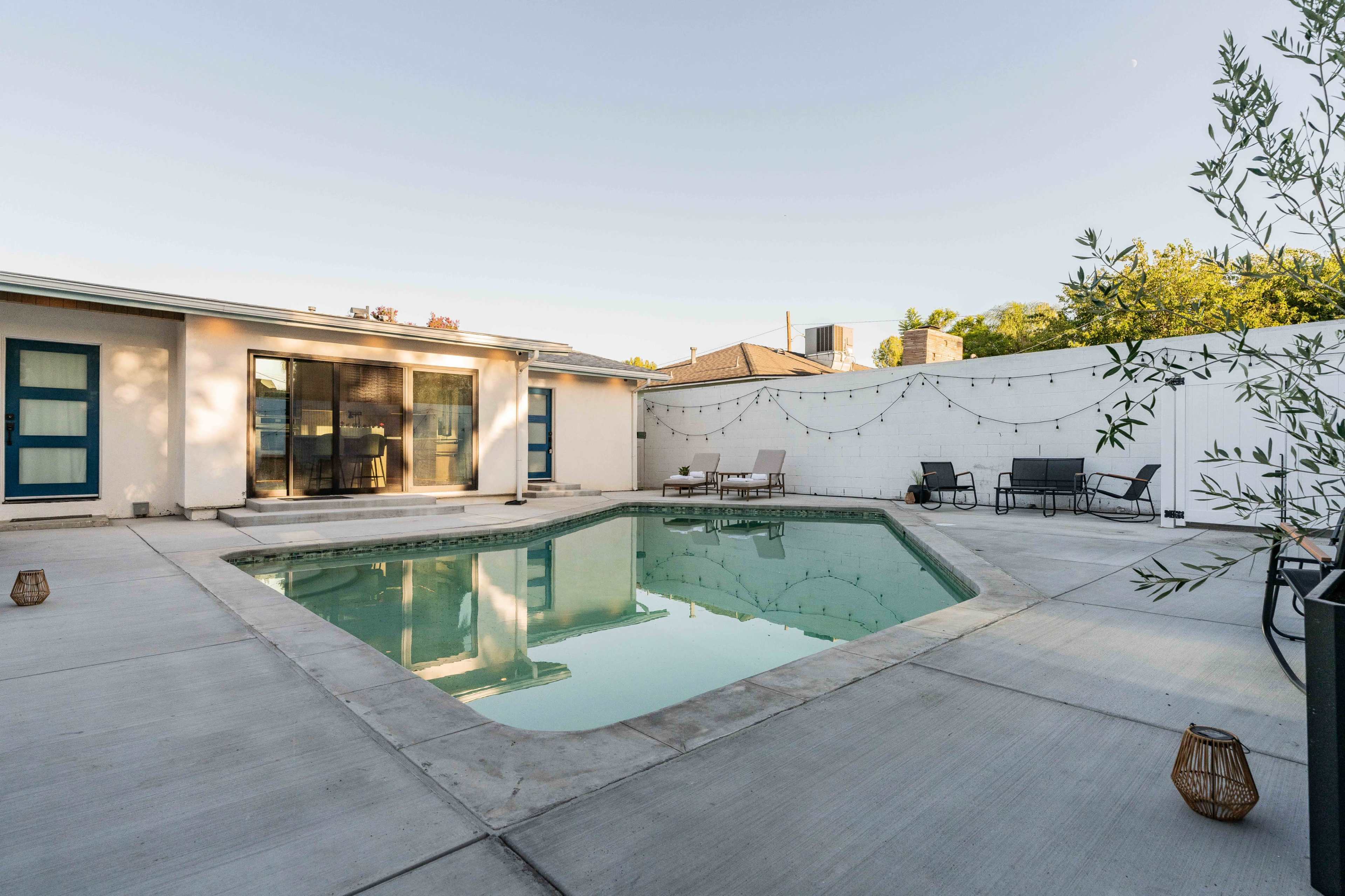 The image shows a rectangular swimming pool surrounded by a concrete patio, with lounge chairs and string lights in a residential backyard.