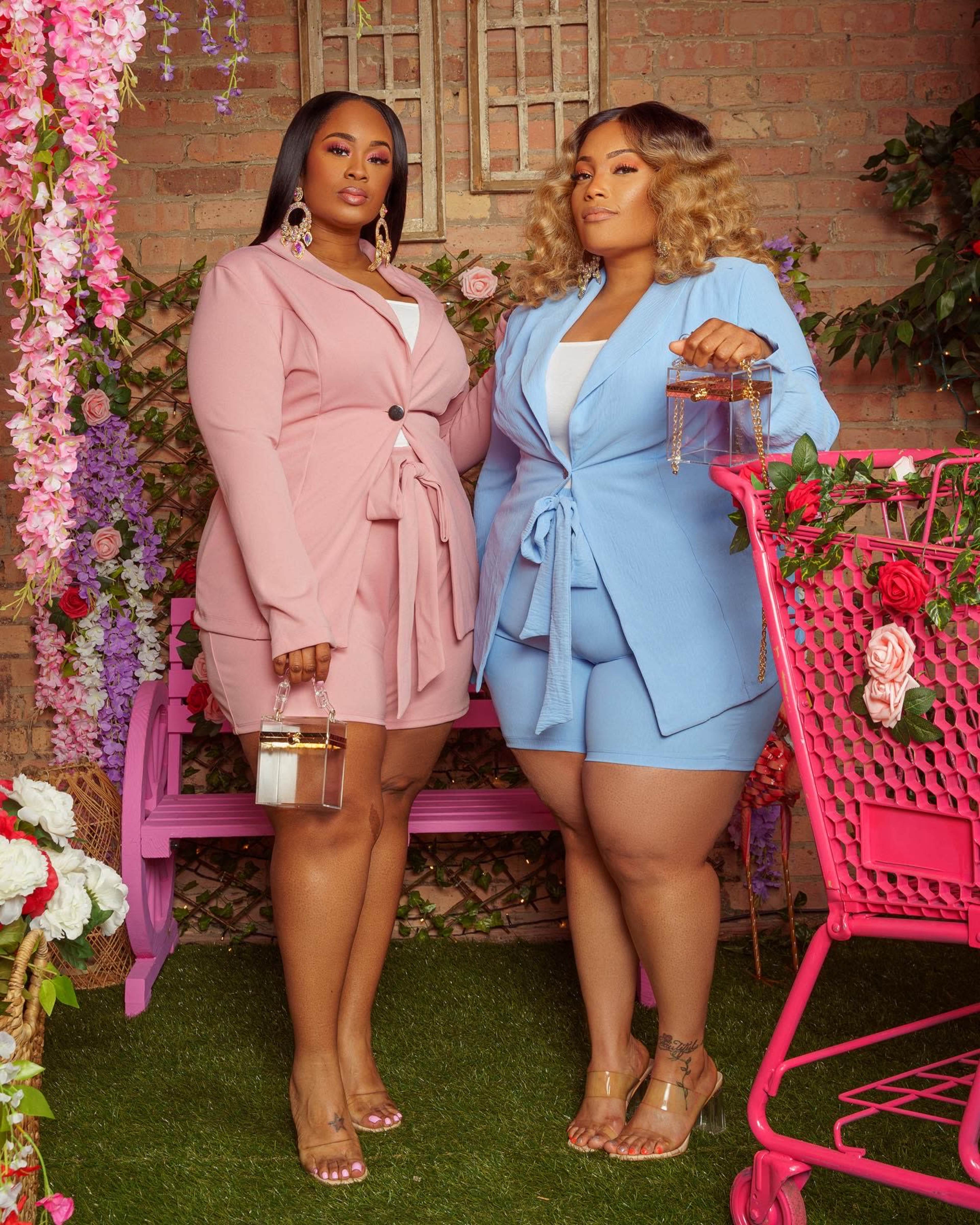Two women stand in a floral setting, each wearing tailored blazers paired with shorts, while one holds a small handbag and the other a purse.