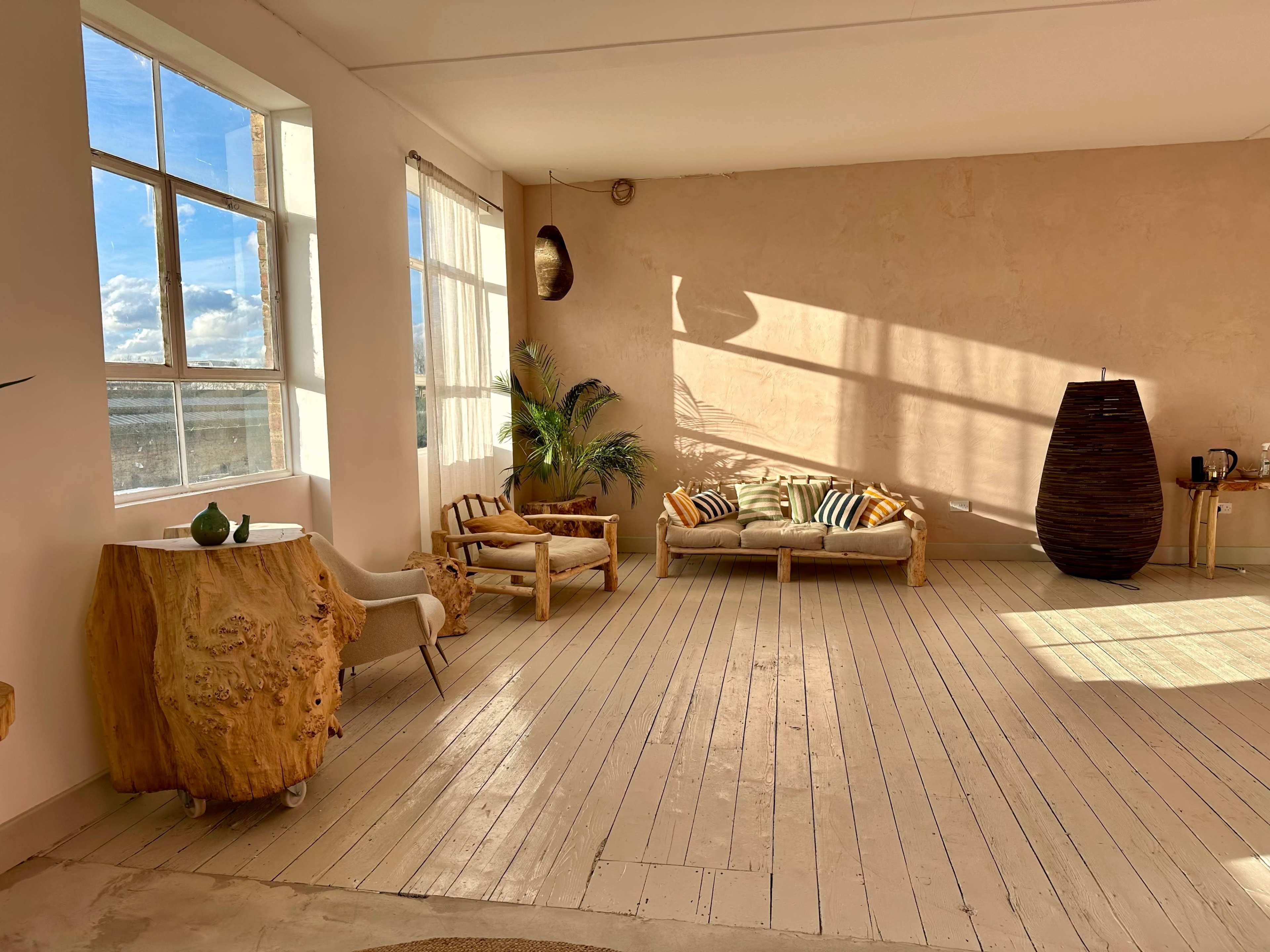 The image shows a bright, minimalist room with large windows, wooden furniture, and a light-colored textured wall.