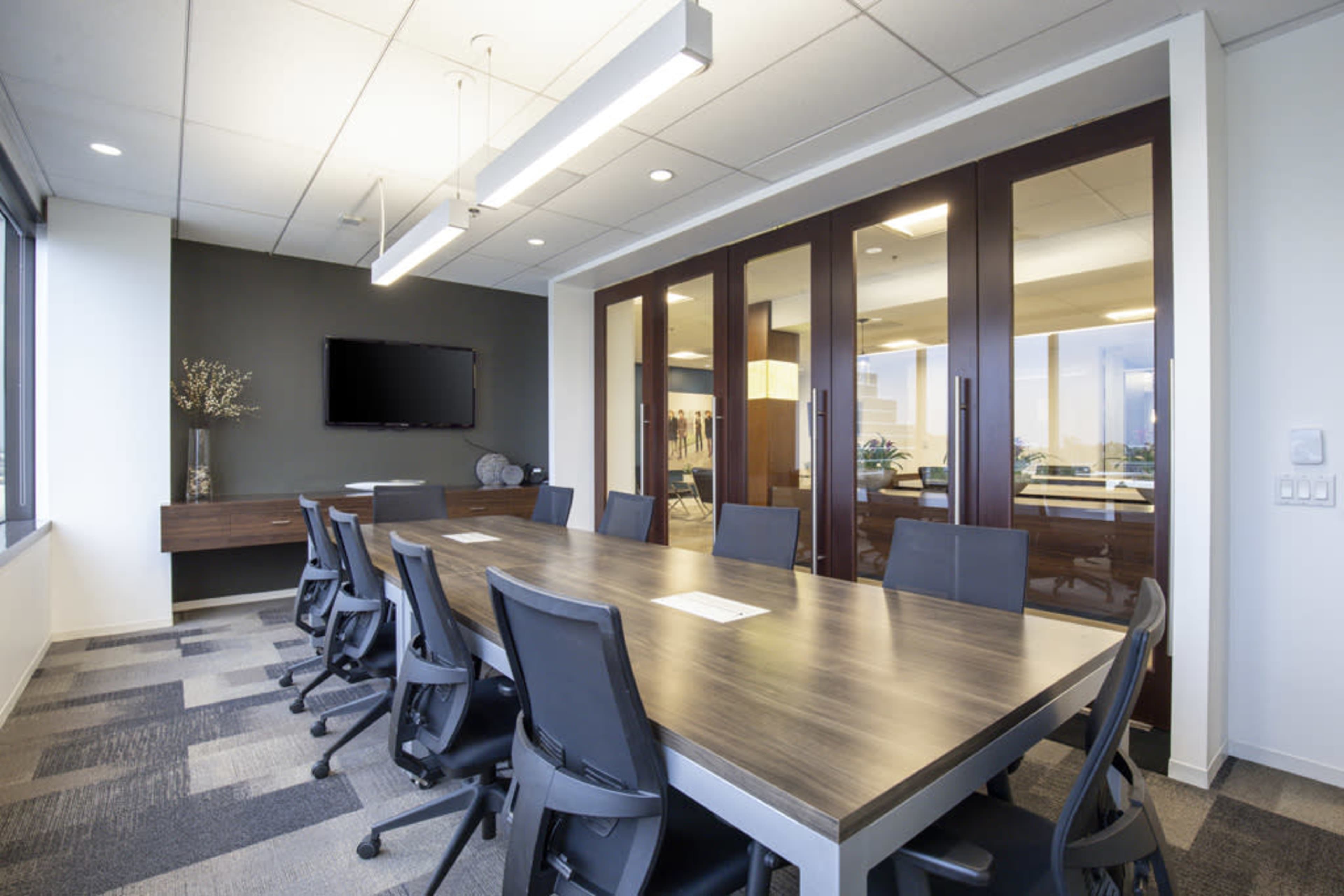A modern conference room features a long wooden table surrounded by ergonomic chairs, with large glass doors leading to another office space.