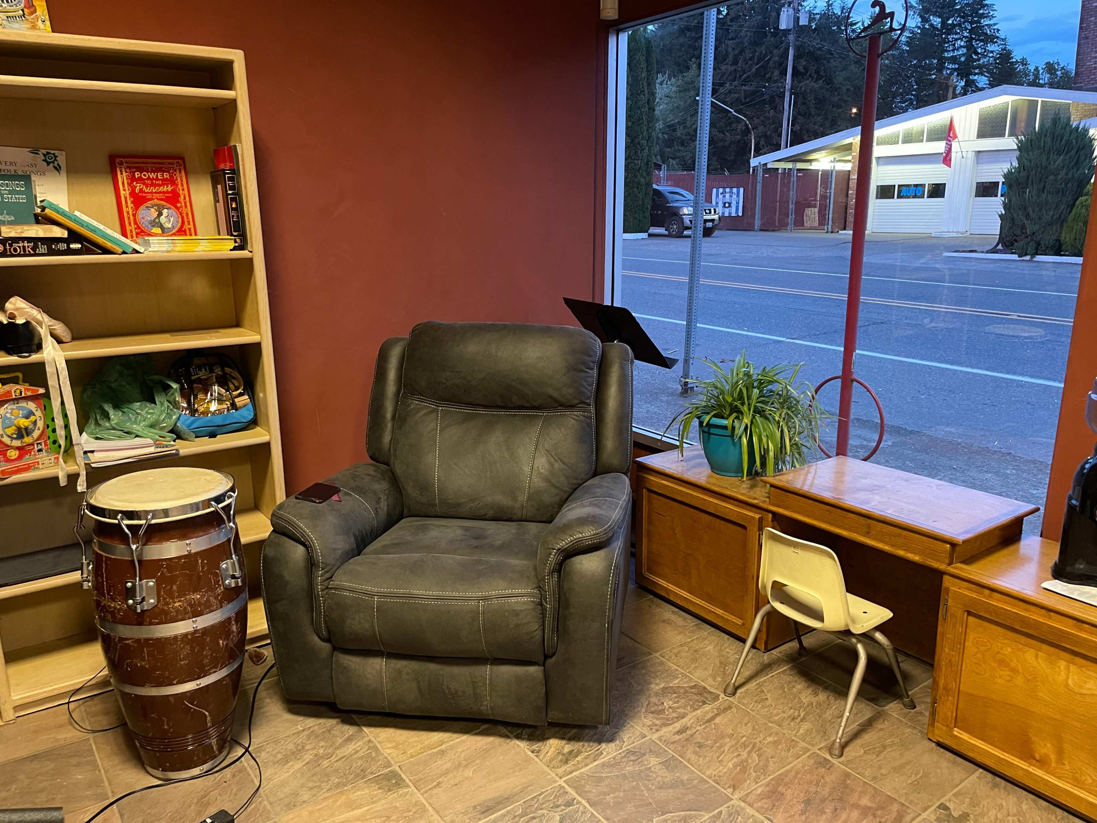 A gray recliner is positioned next to a wooden desk with a small chair, alongside a conga drum and a potted plant, within a room that has large windows overlooking a street.