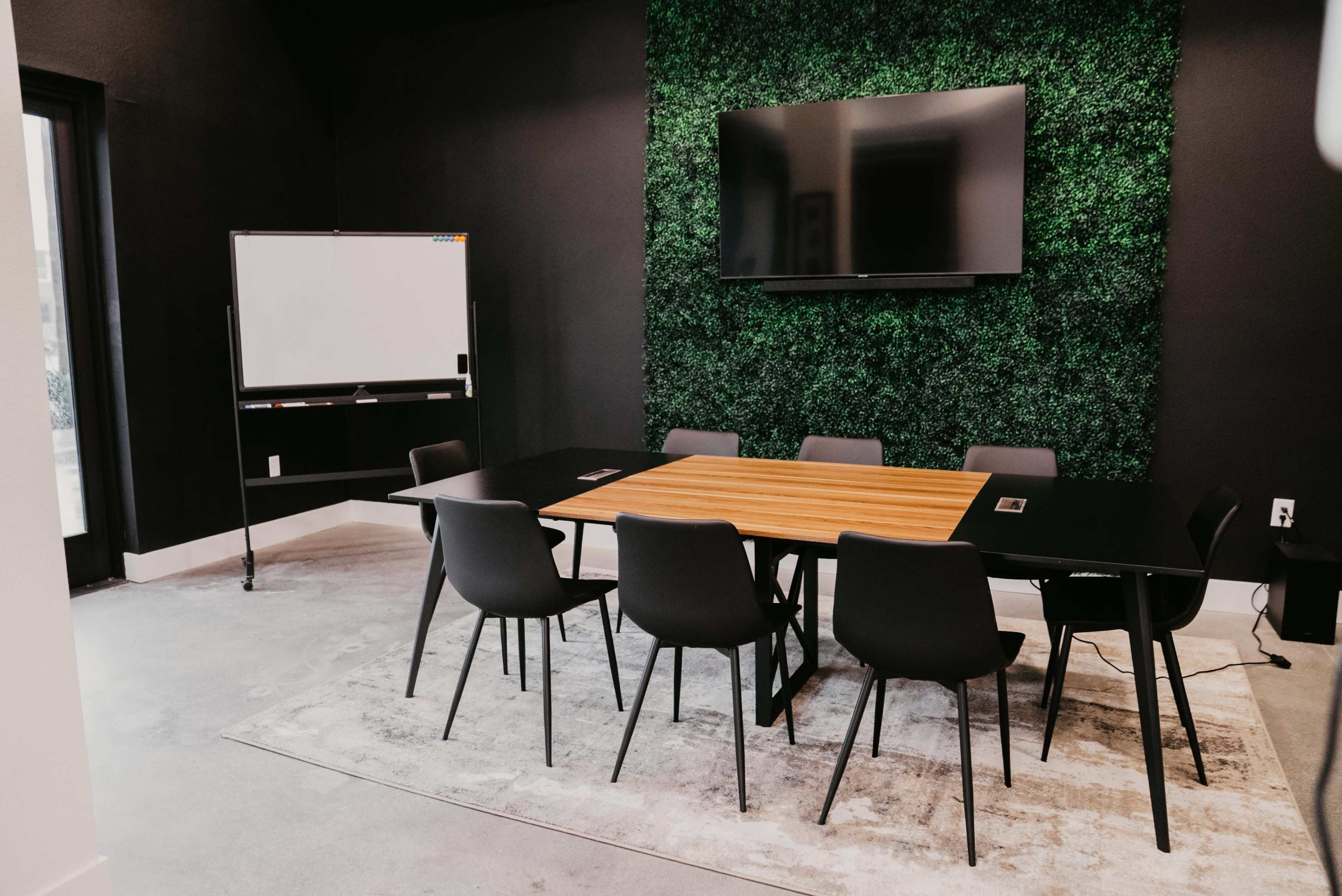 A modern conference room features a wooden table surrounded by chairs, green wall accents, a television mounted on the wall, and a whiteboard nearby.