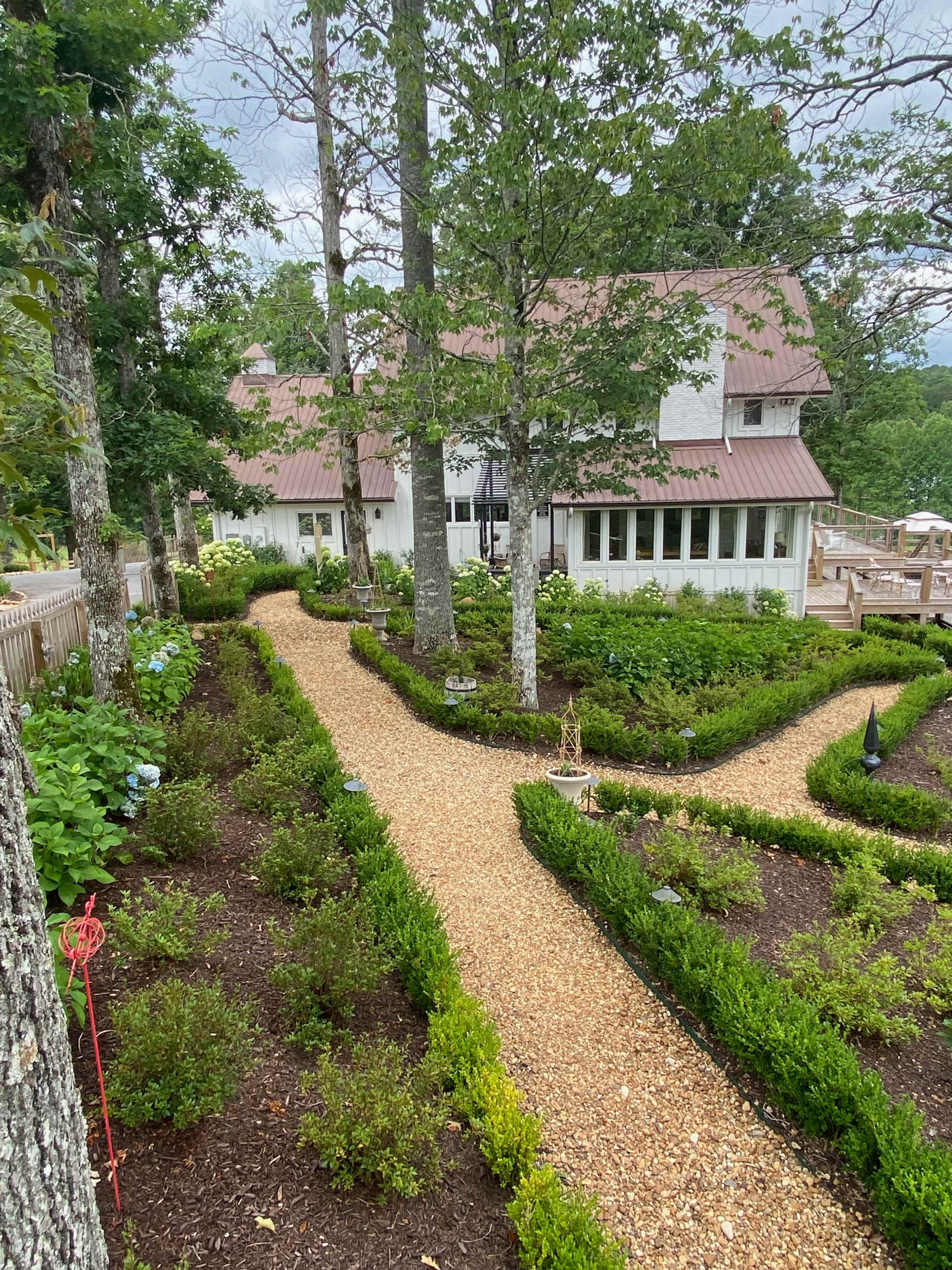 A path of gravel winds through a landscaped garden with neatly trimmed hedges and trees, leading to a white house with a metal roof.