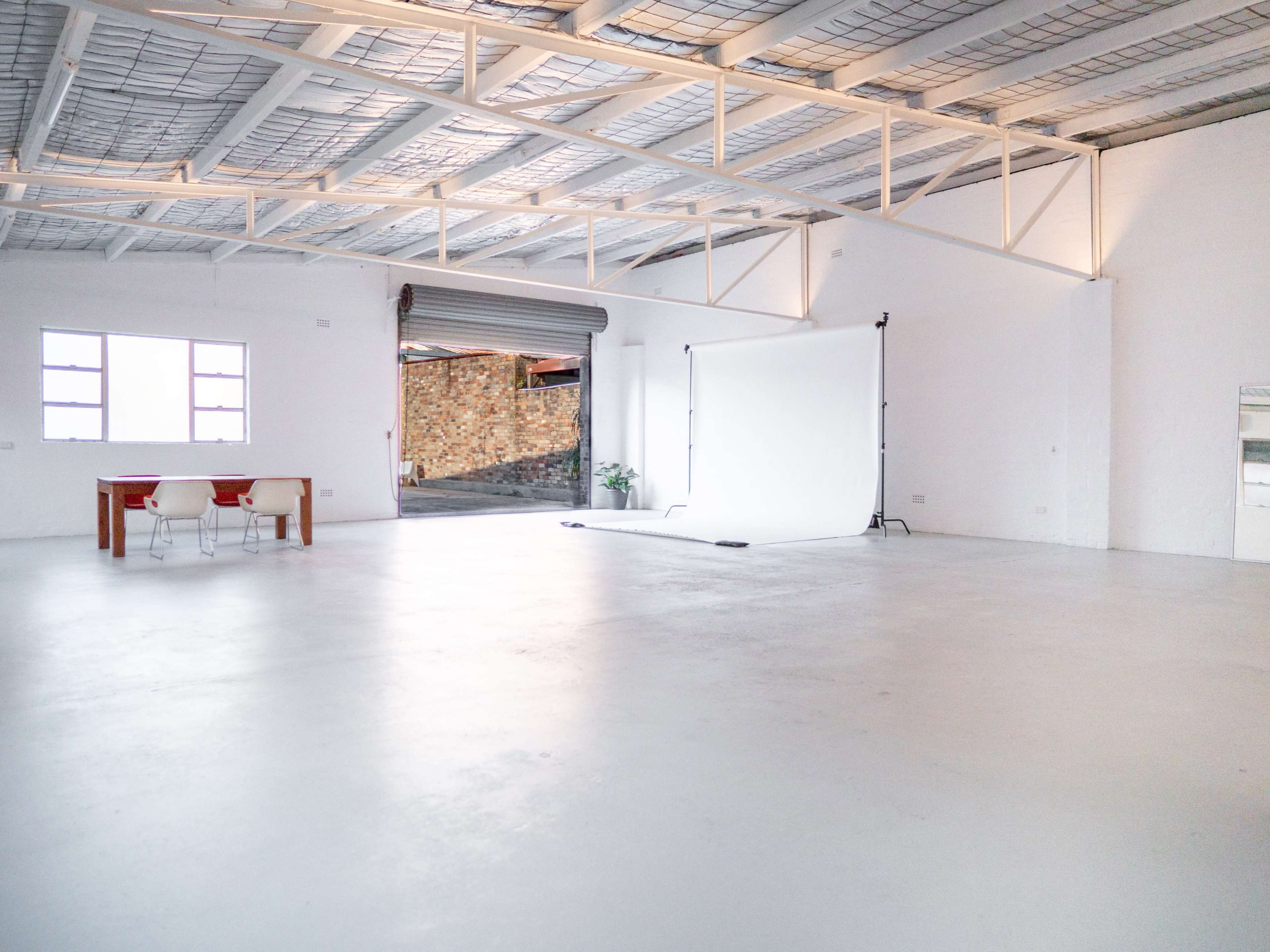A spacious, empty industrial-style room with a white backdrop set up, a table, and two chairs.