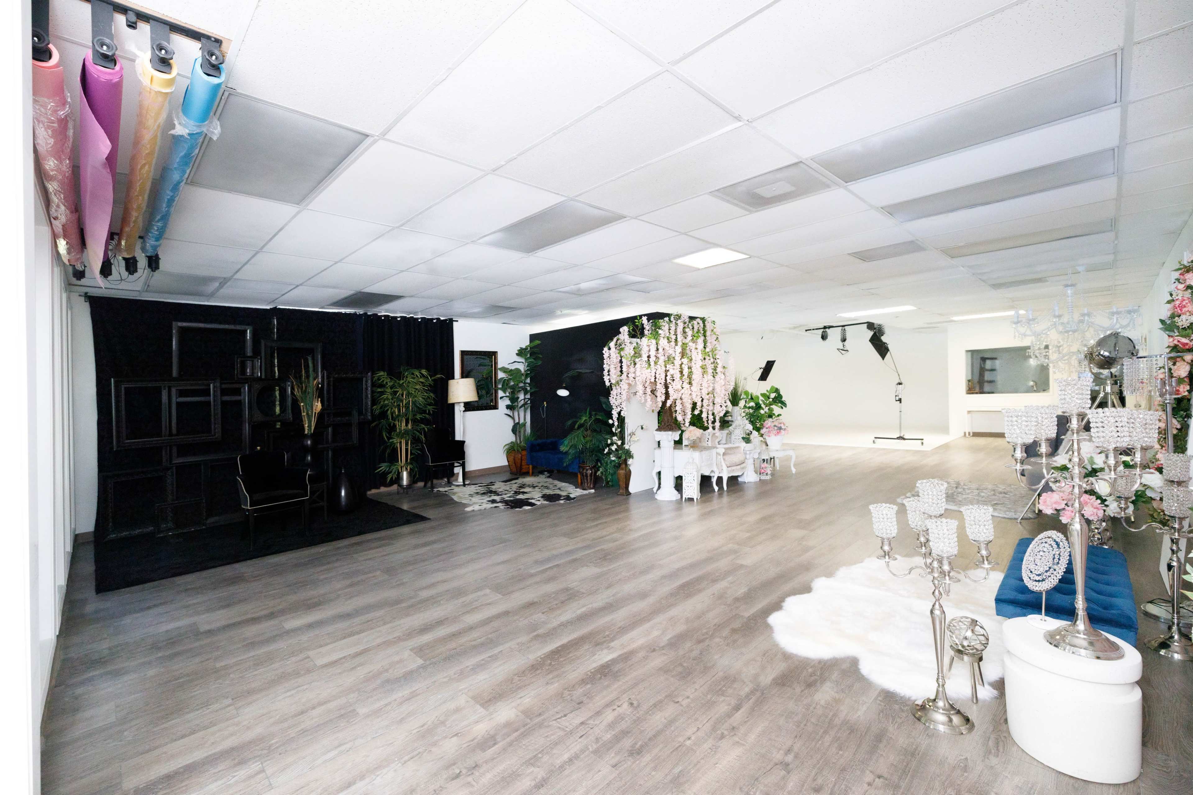 The image shows a spacious interior room with wooden flooring, featuring different styled seating areas, decorative plants, and hanging floral arrangements.