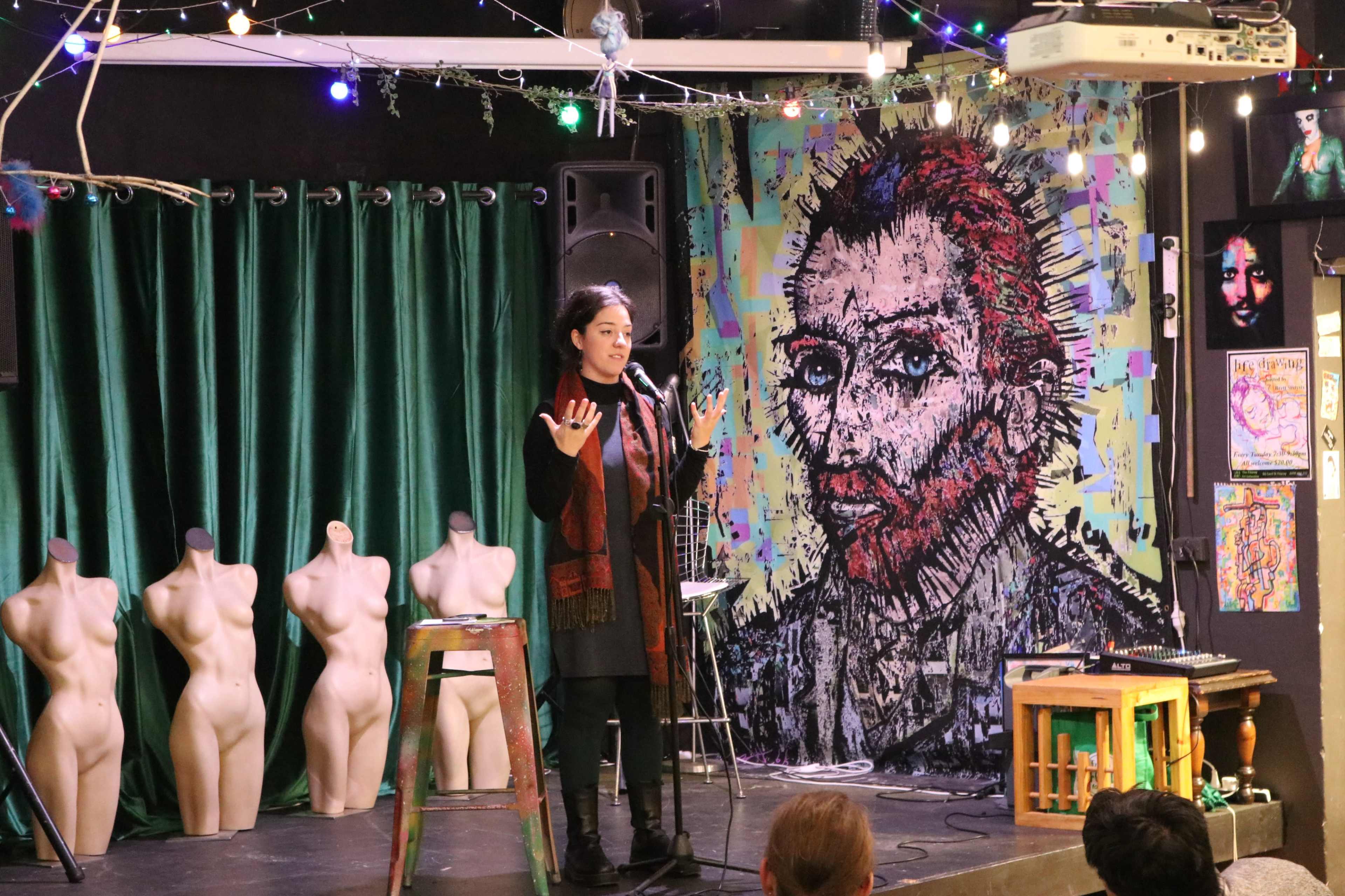 A speaker stands on a stage in front of a colorful mural resembling a famous artist, with green curtains and artistic mannequins in the background.