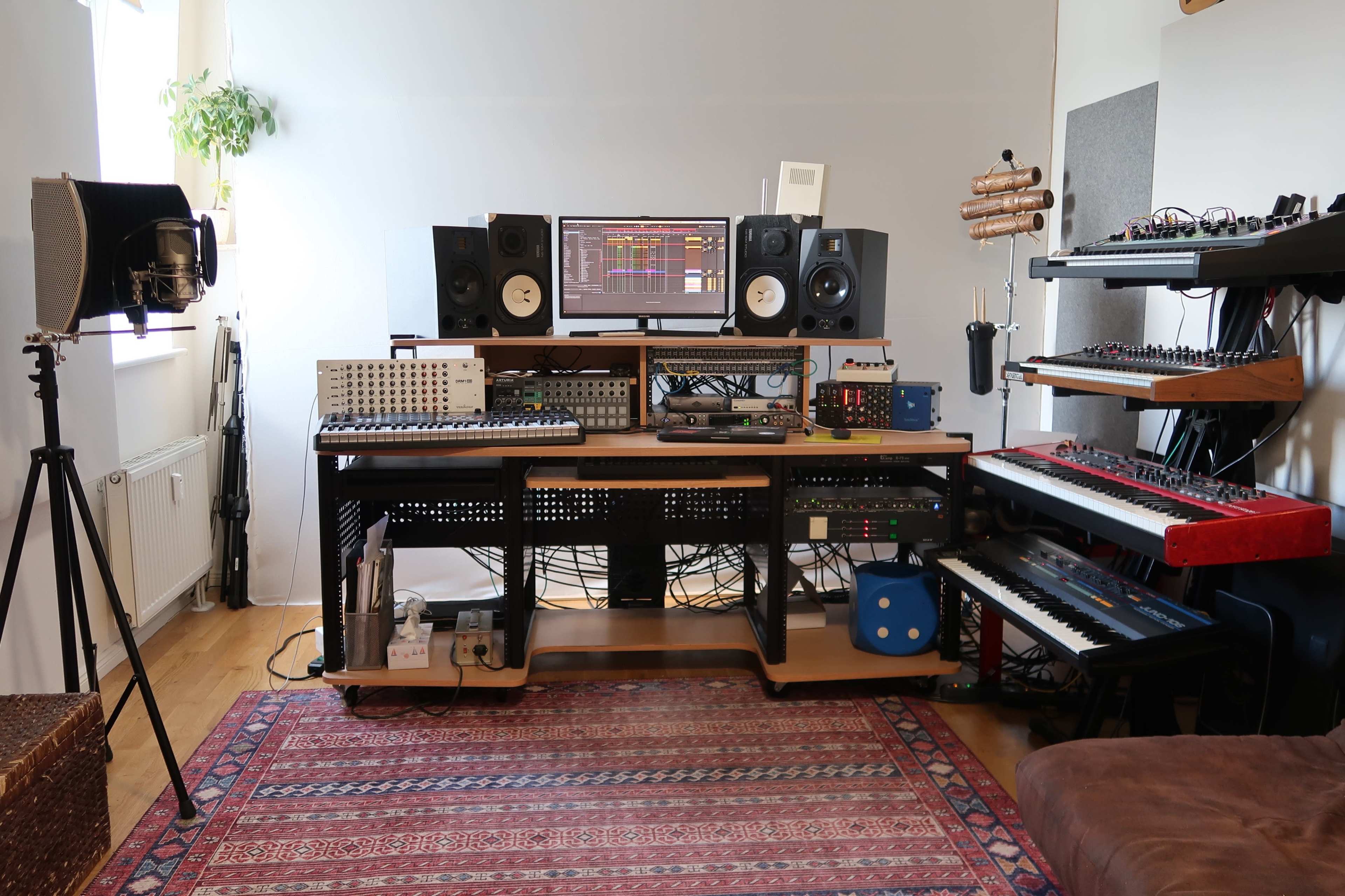 The image shows a music production studio featuring a computer monitor, audio speakers, synthesizers, a mixing console, and various electronic instruments arranged on a wooden desk.