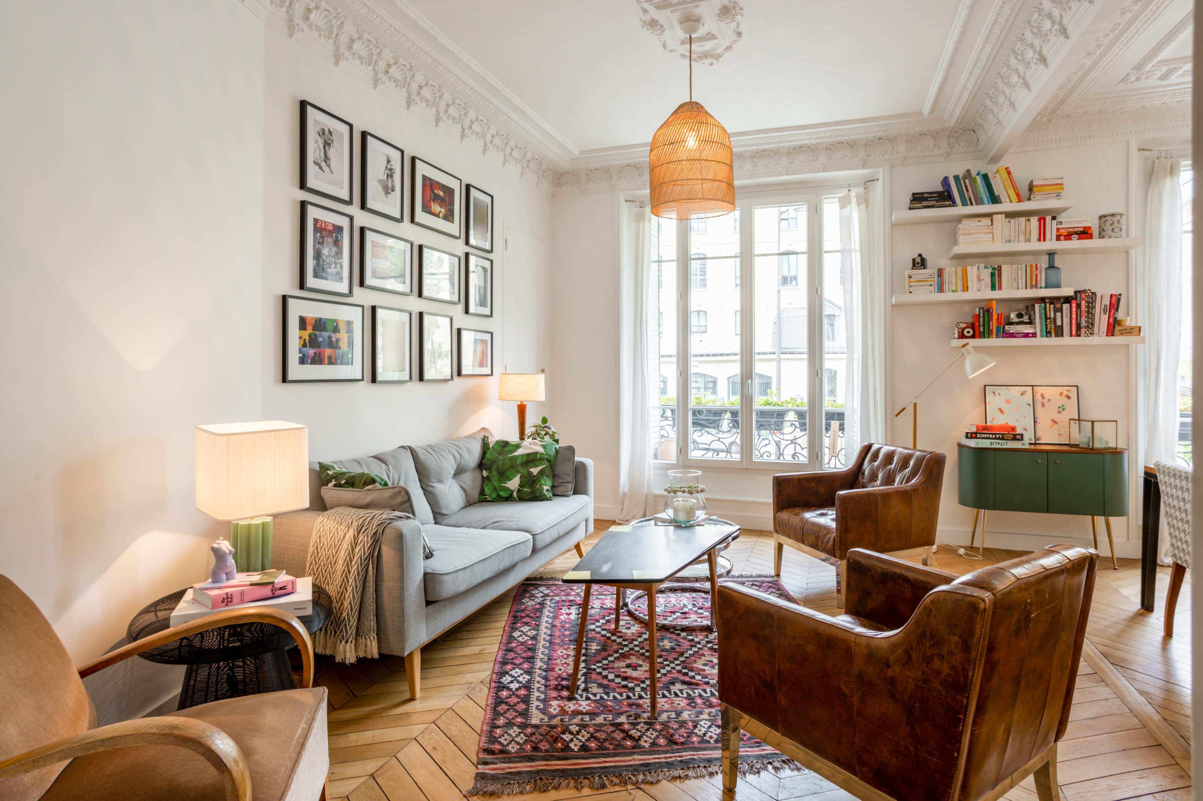 A cozy living room features a gray sofa, wooden armchairs, a coffee table, and bookshelves, with large windows allowing natural light.