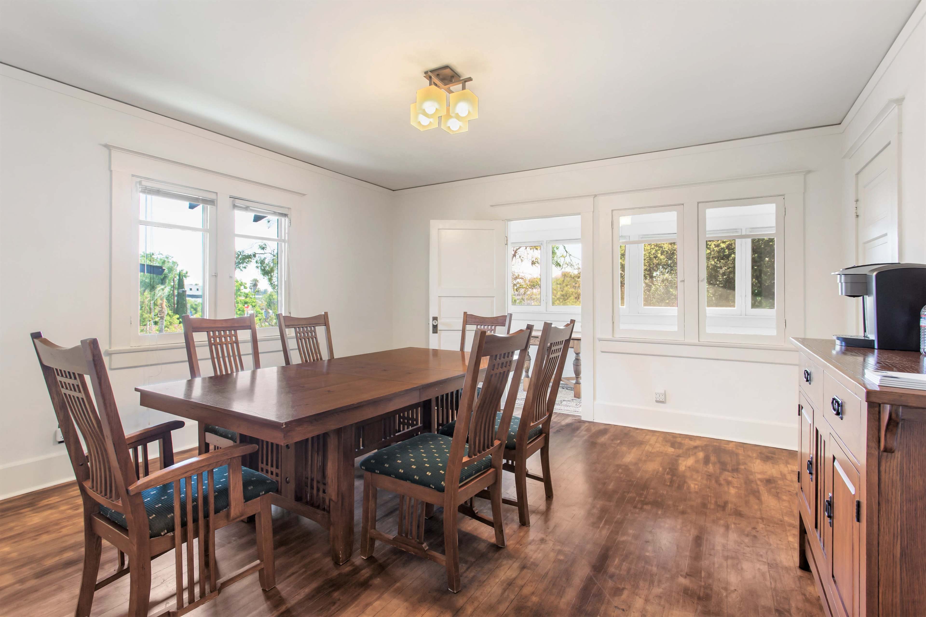 A wooden dining table surrounded by eight chairs is positioned in a well-lit room with large windows and a sideboard.