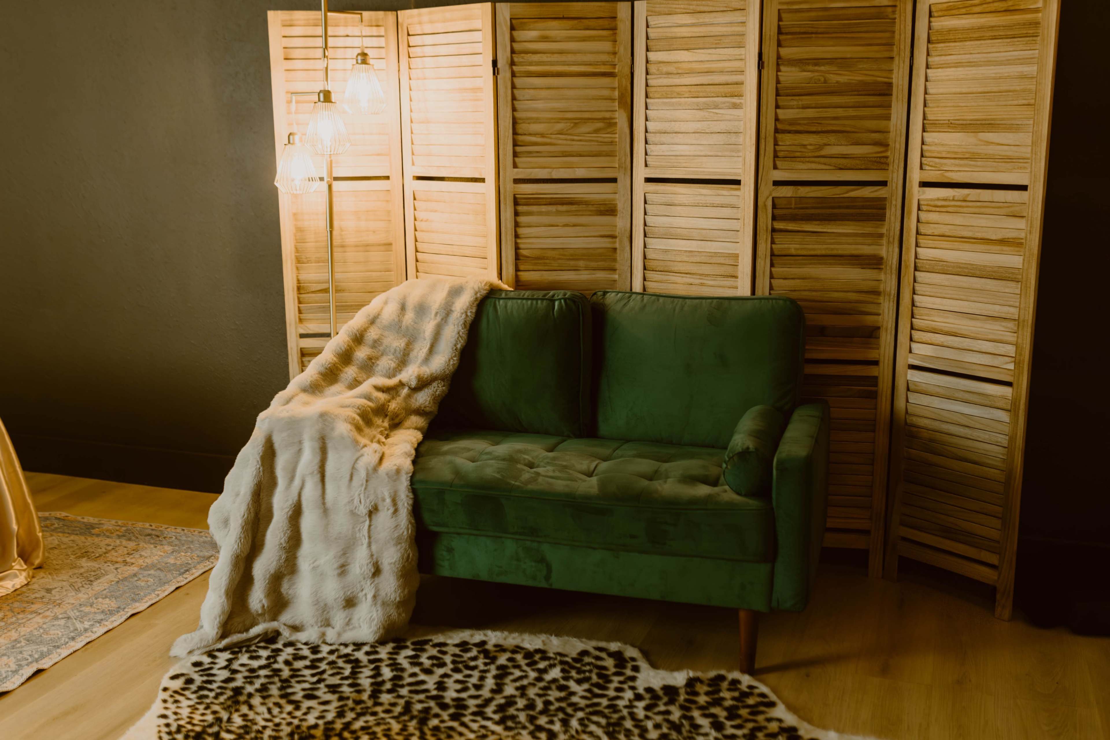 A green velvet couch is draped with a light-colored throw blanket, placed next to a wooden screen and illuminated by pendant lights.