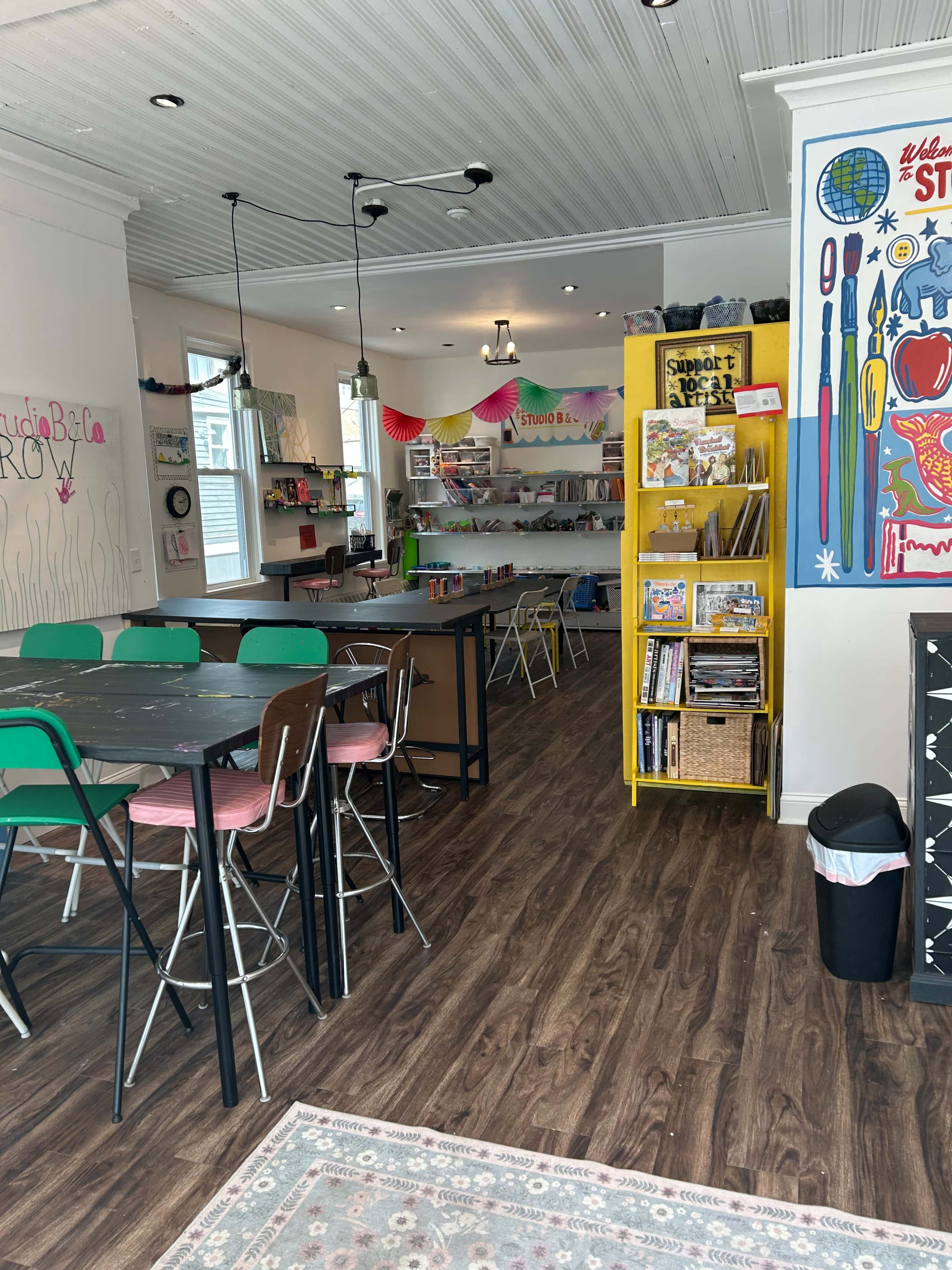 The image shows a bright, open creative workspace with tables and chairs arranged for group activities, shelves filled with books and supplies, and colorful decorations on the walls.