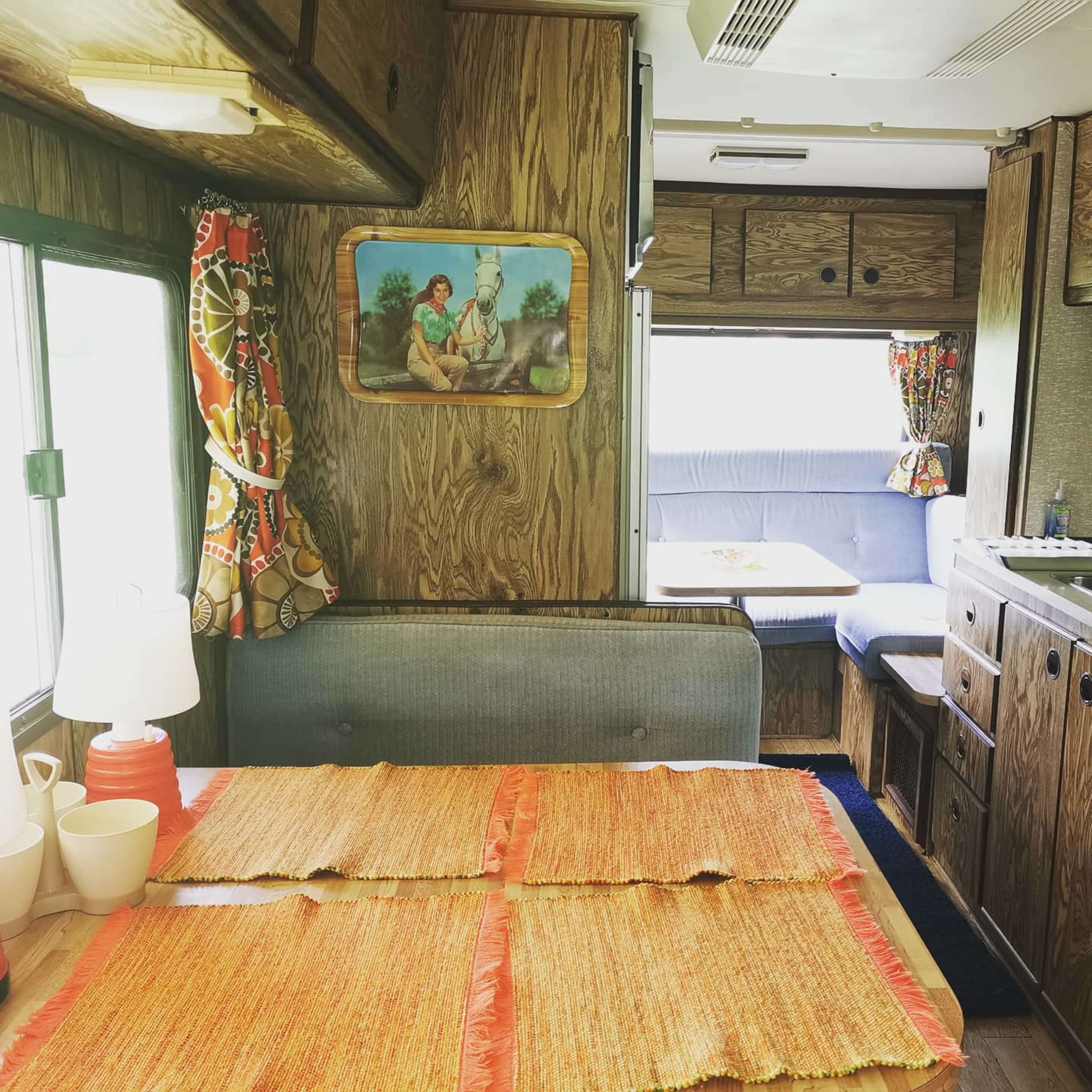 The interior of a vintage camper features a dining area with a table covered in a woven mat, a lamp, and a wall-mounted painting, alongside a seating area with a fold-out bed in the background.