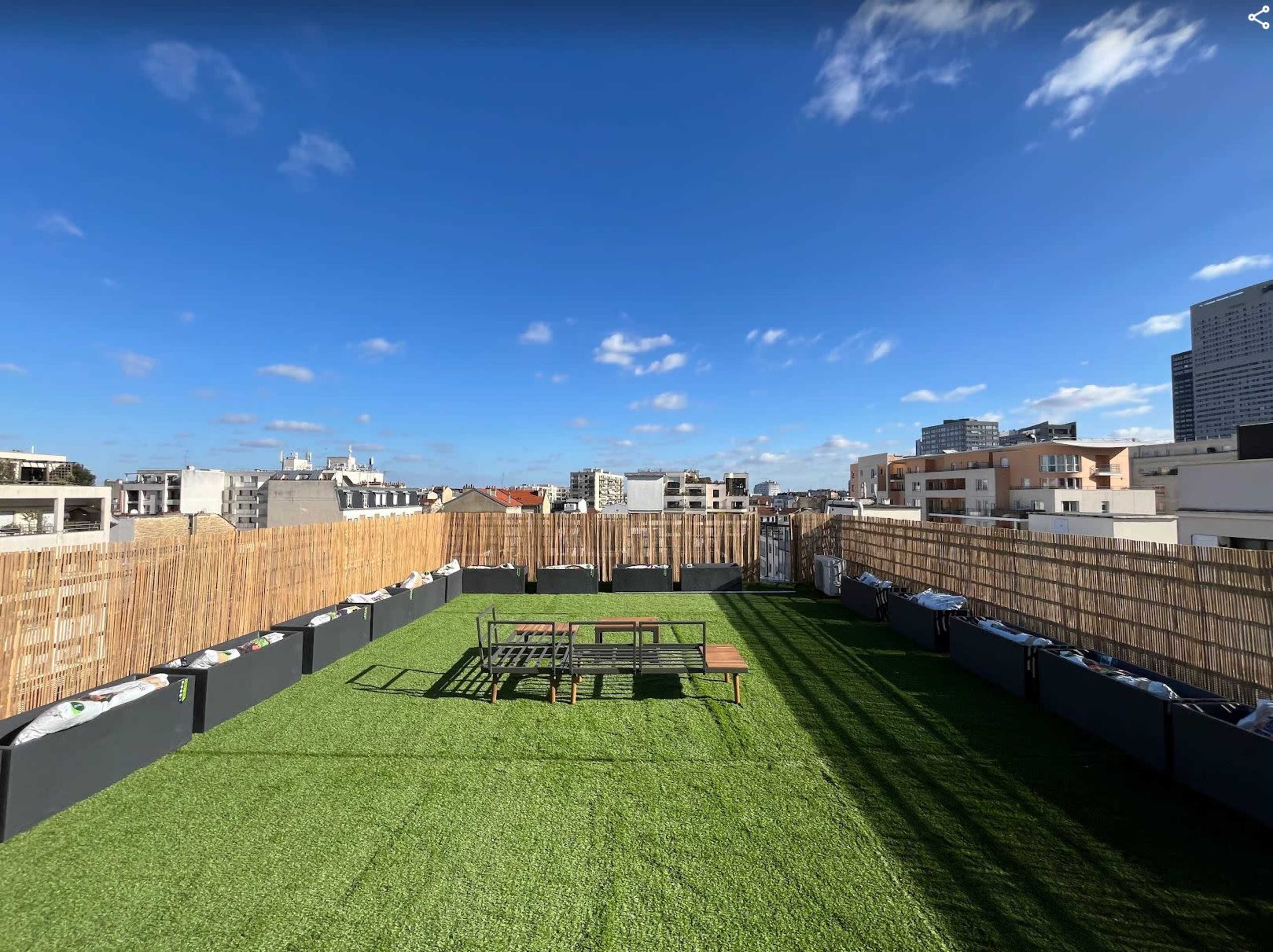 The image shows a rooftop space with artificial grass, wooden picnic tables, and a bamboo fence surrounding the area under a clear blue sky.