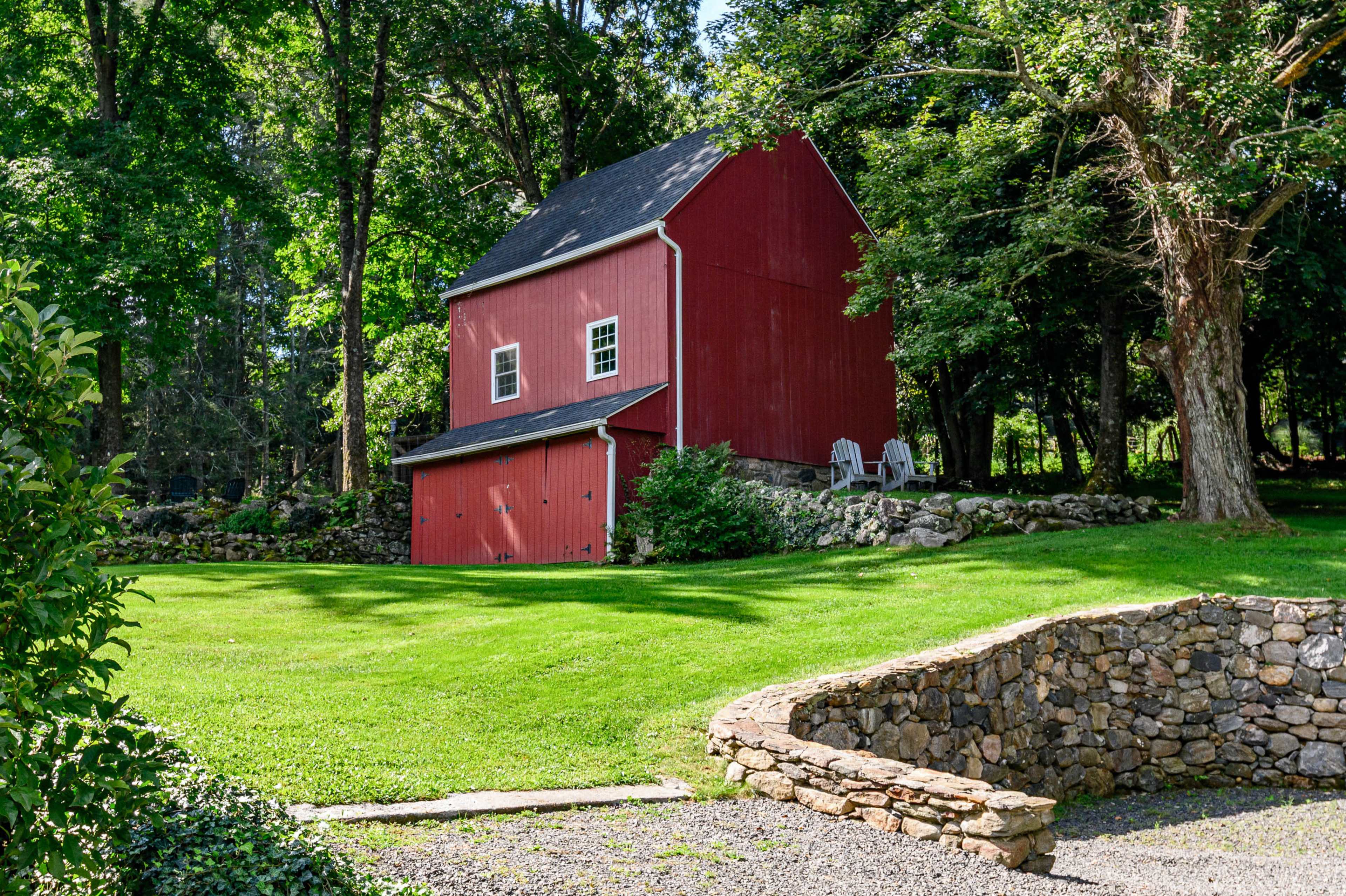 A red barn stands in a green yard surrounded by trees, with a stone wall and two chairs visible nearby.