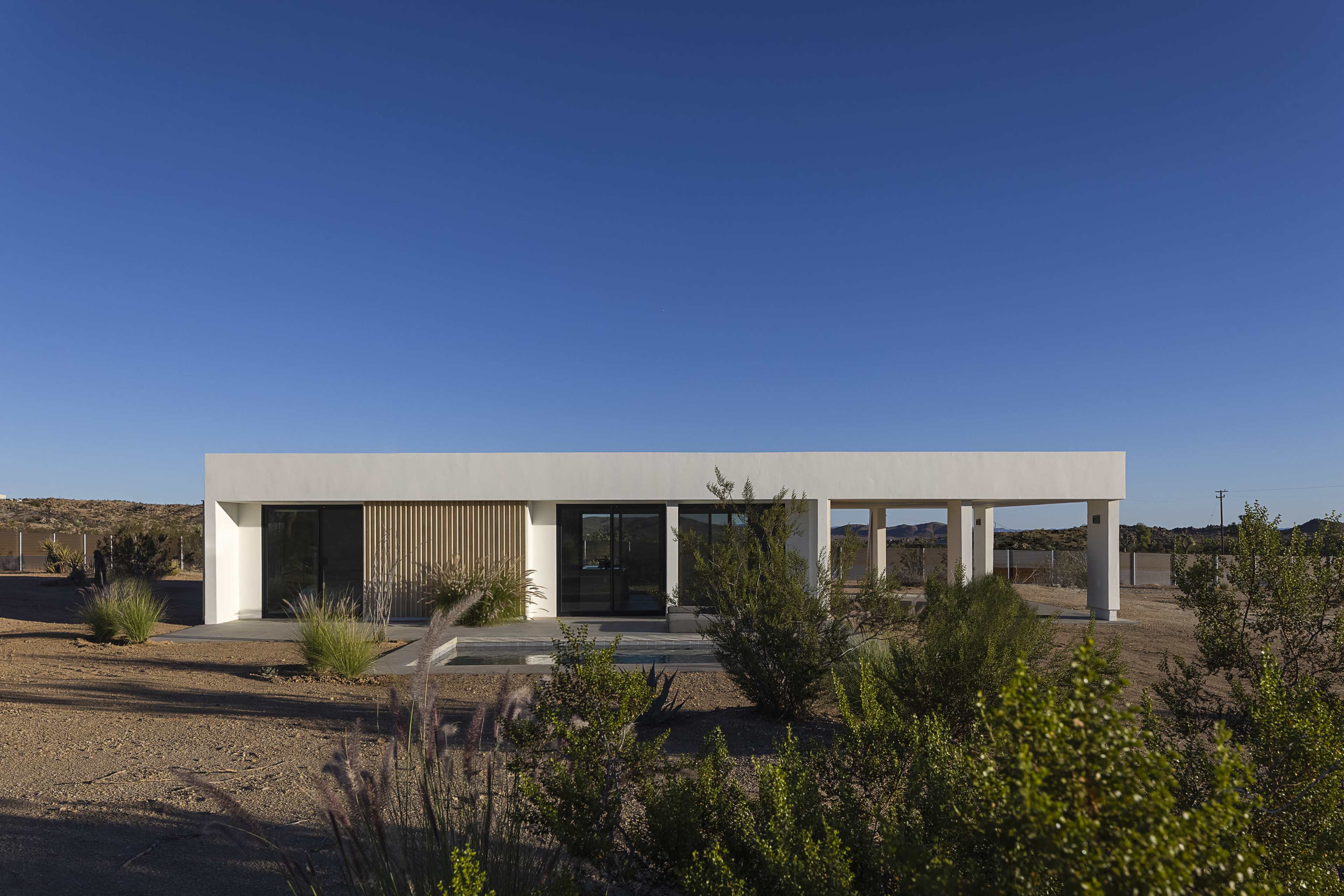 A modern, single-story house with a white exterior and wooden accents is set against a clear blue sky in a desert landscape.