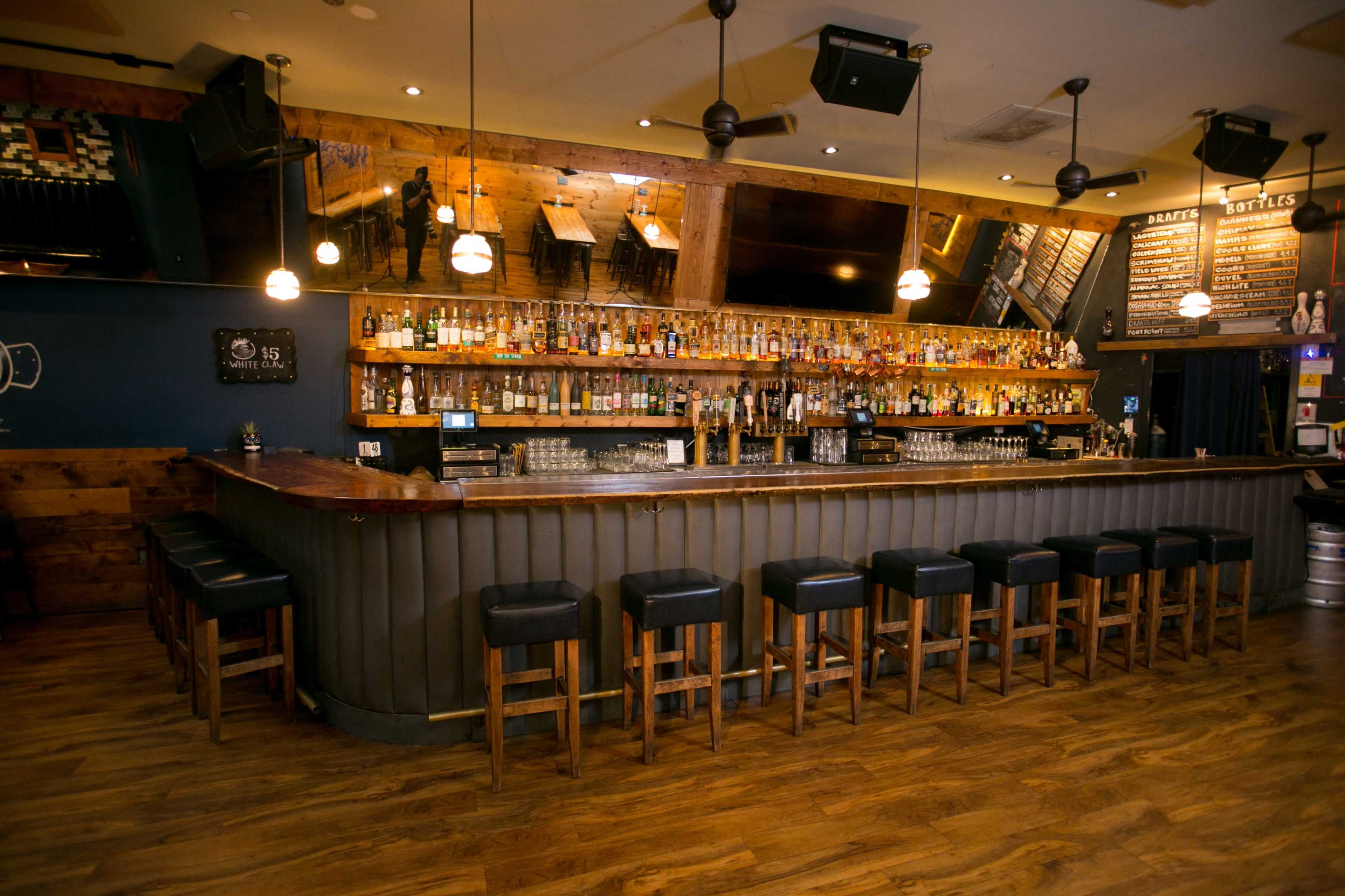 The interior of a bar features a long wooden countertop with rows of liquor bottles behind it and several bar stools lined up in front.
