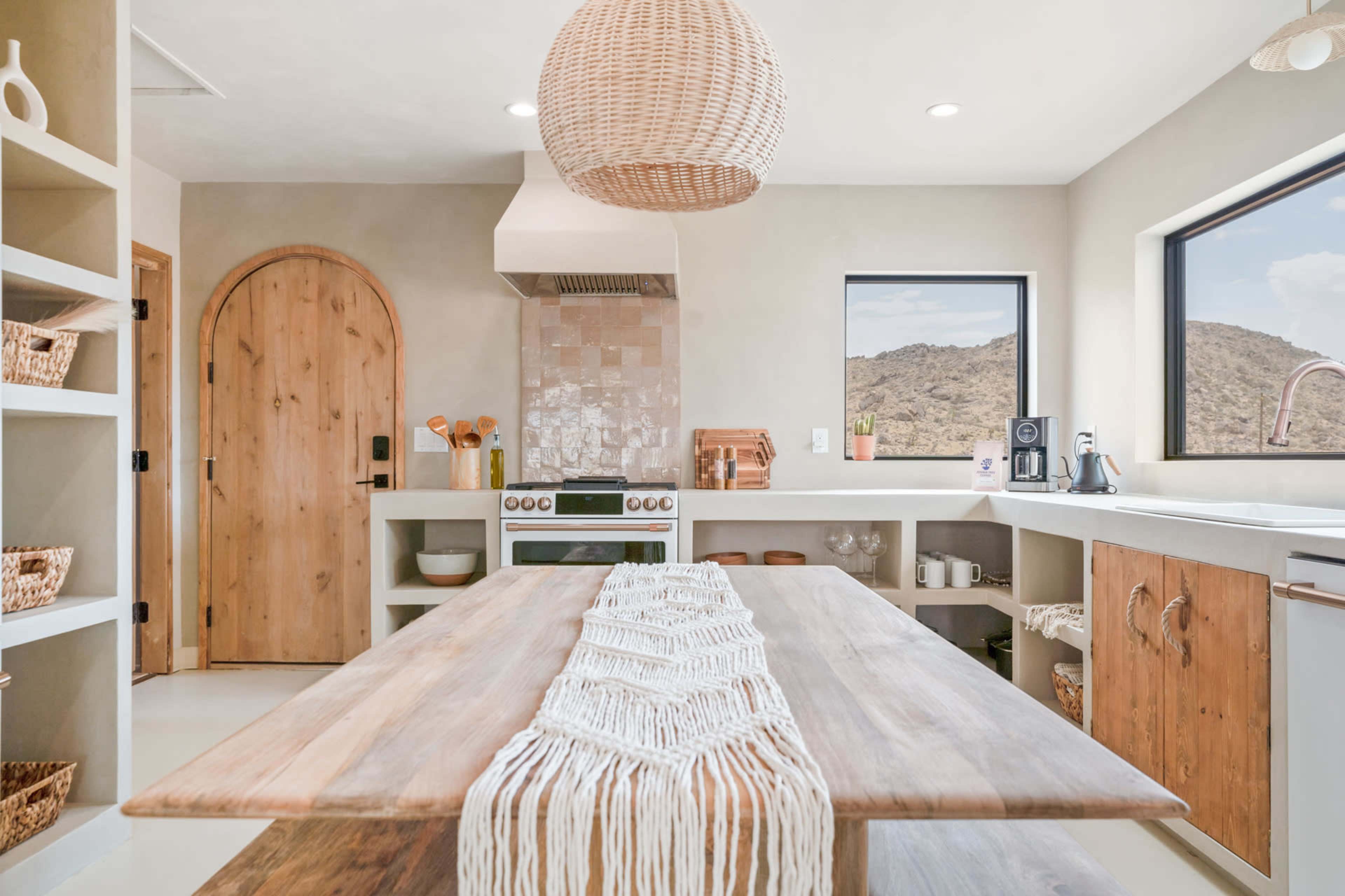A modern kitchen features a wooden dining table, a round pendant light, and large windows with desert views.