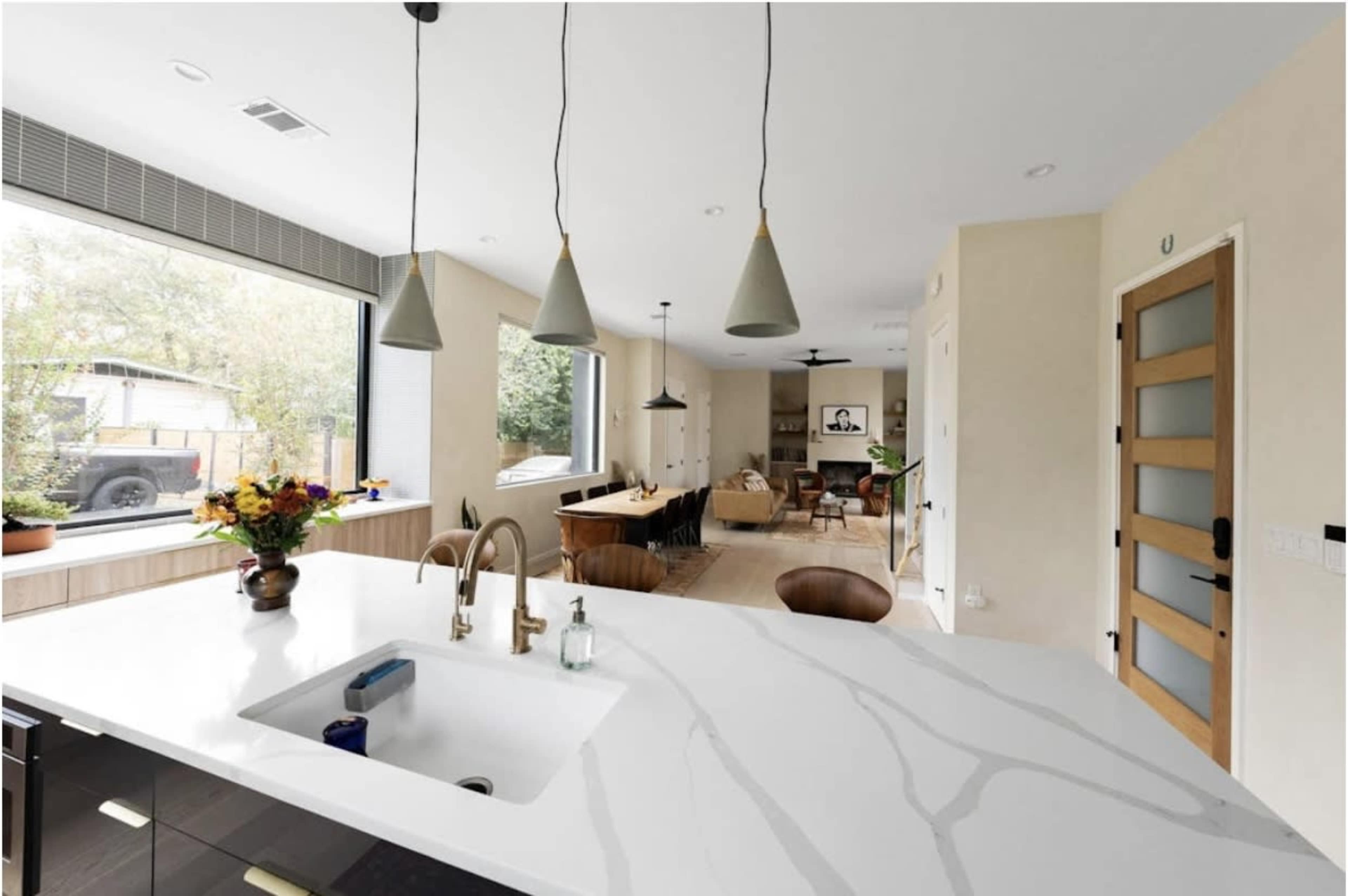 A modern kitchen with white marble countertops features a sink and gold faucets, while a dining area with wooden furniture is visible through large windows.