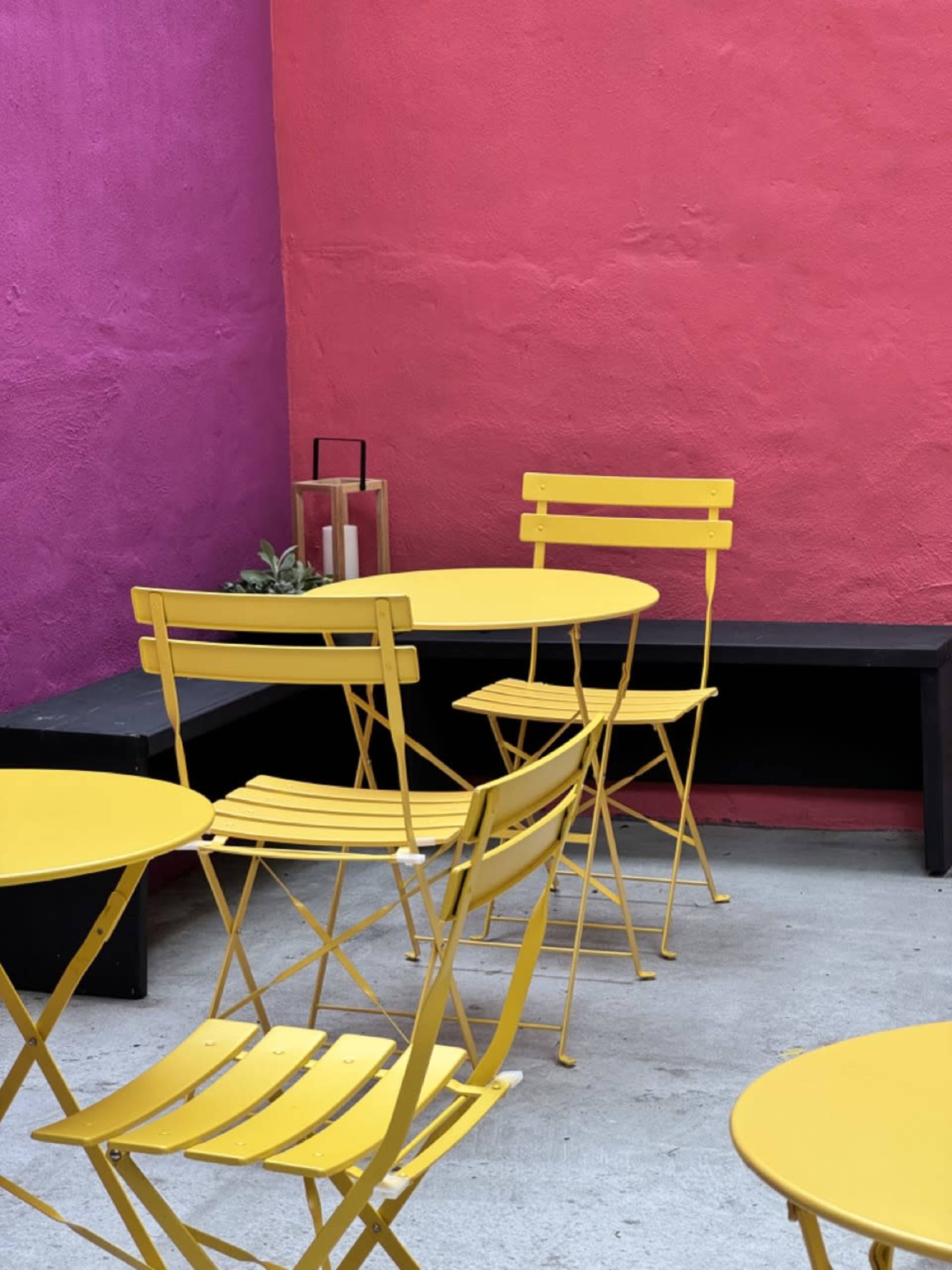 The scene features yellow metal tables and chairs arranged in front of a vibrant pink wall.