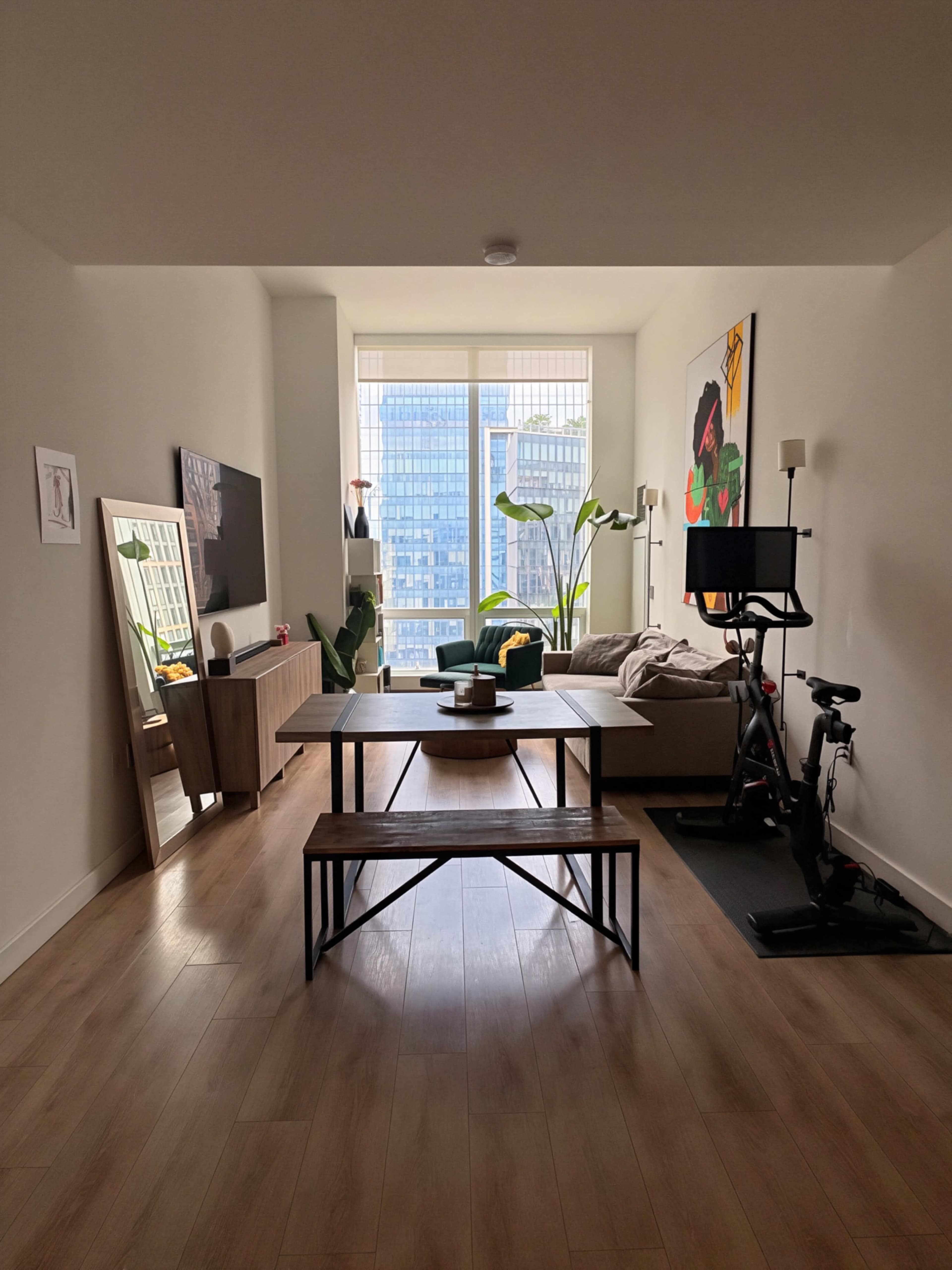 The image shows a modern living room with a minimalist design, featuring a coffee table, a couch, a mirror, a television, and a stationary bike.
