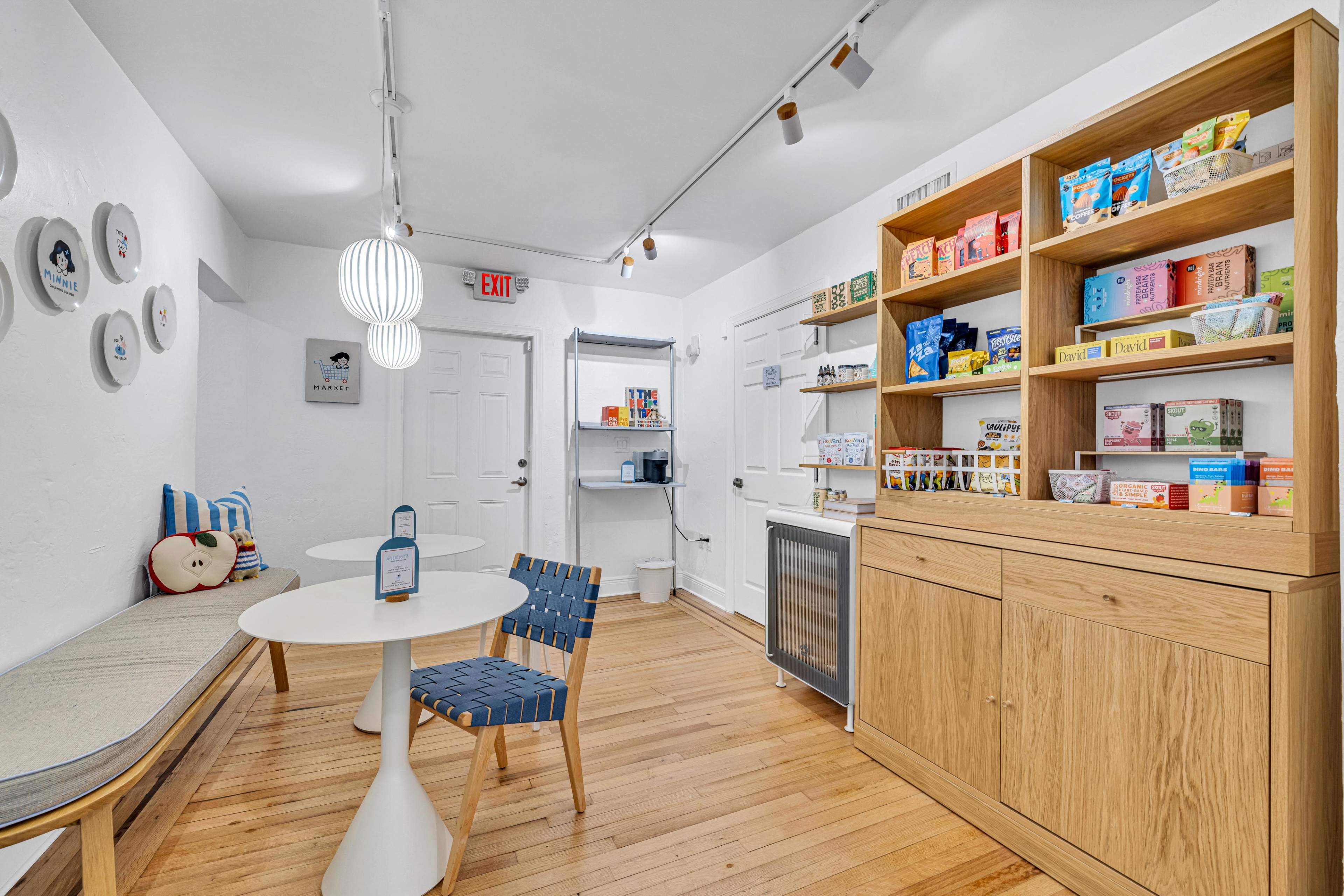 The image shows a small room with a table and chairs, a bench along one wall, and shelves filled with various packaged items alongside a kitchenette area.