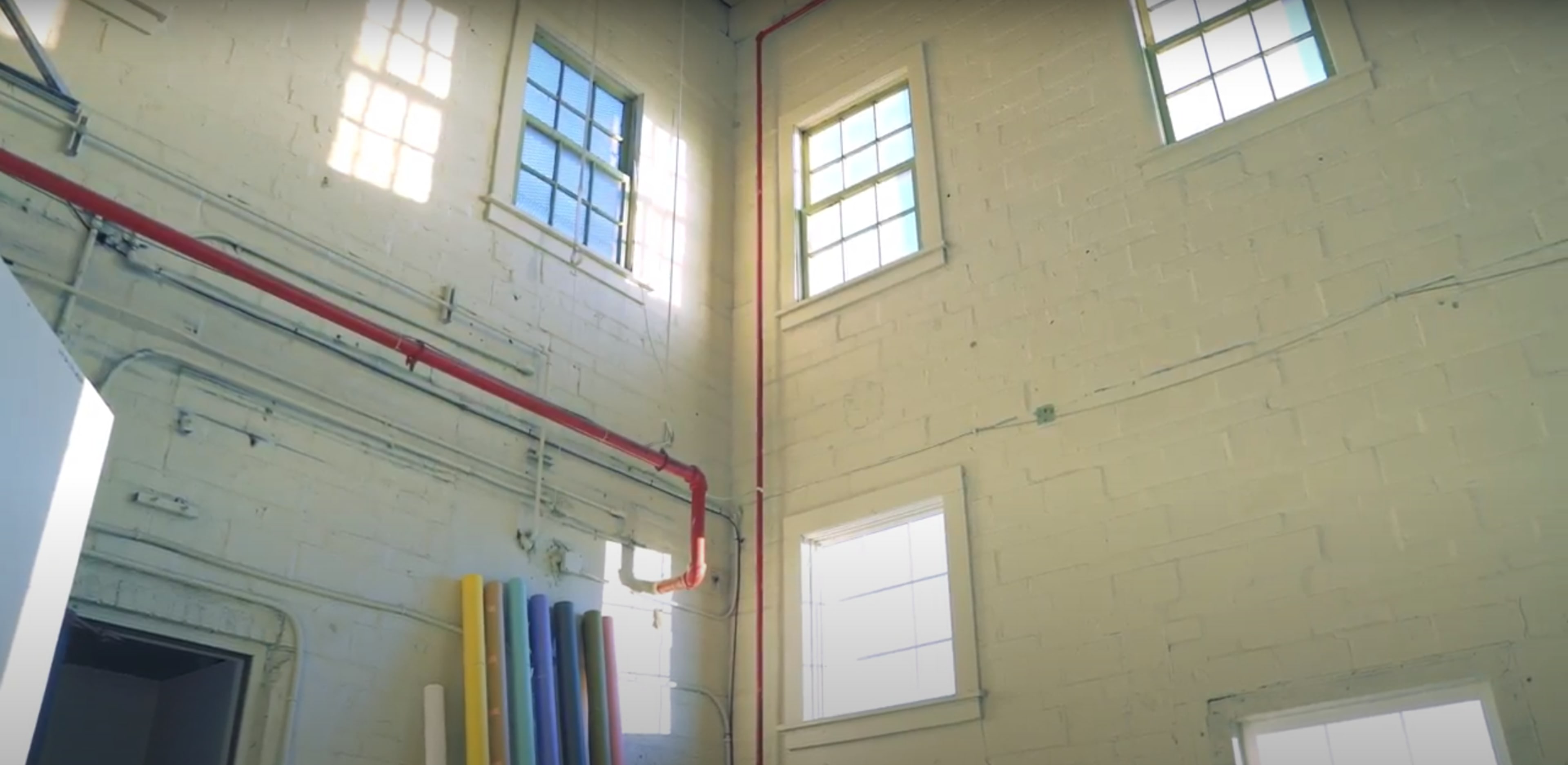 The interior of a room with high ceilings, featuring several windows and colorful pipes leaning against the wall.