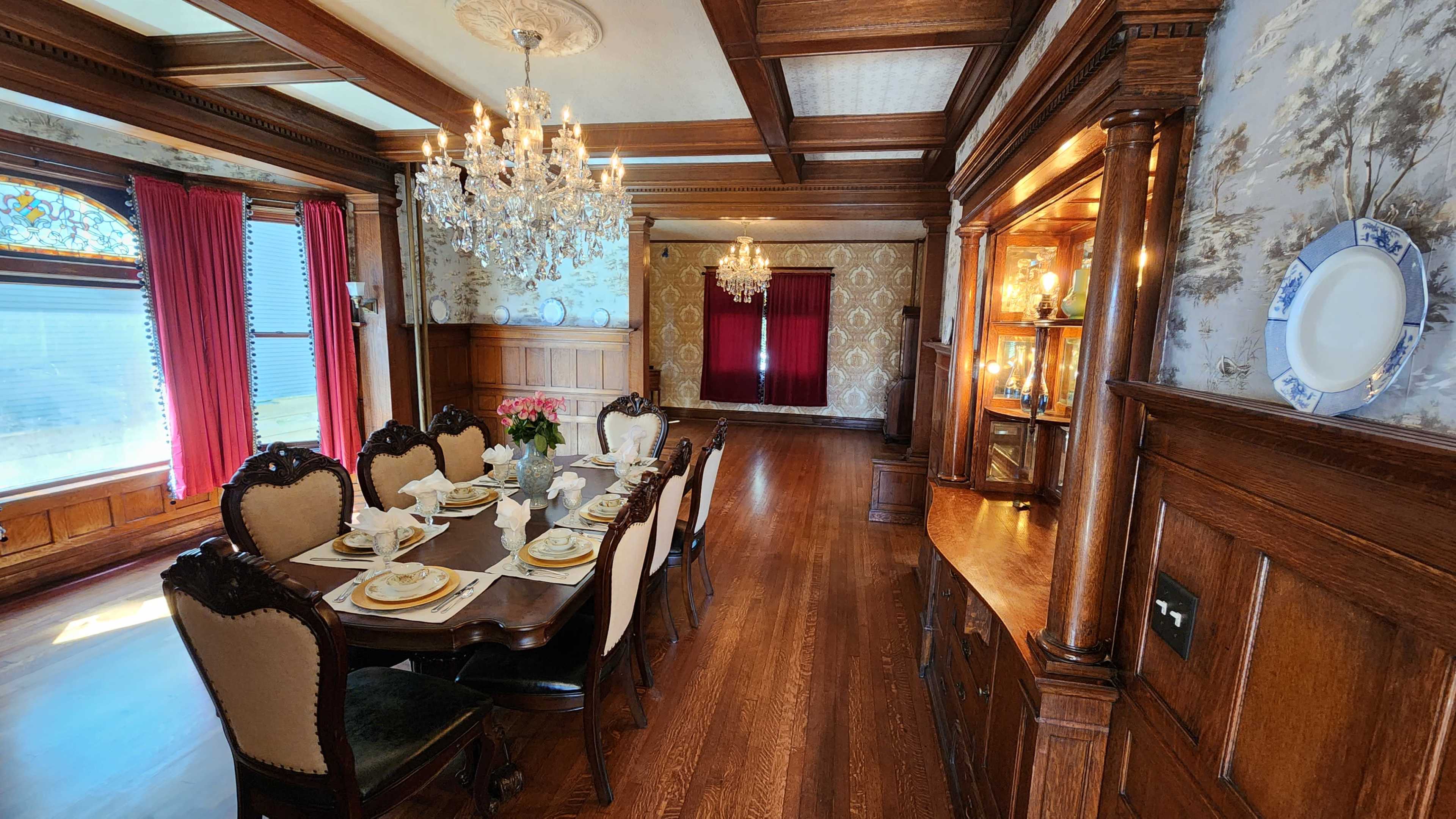 A formal dining room with a large wooden table set for a meal, surrounded by high-backed chairs, and adorned with chandeliers and decorative wall features.