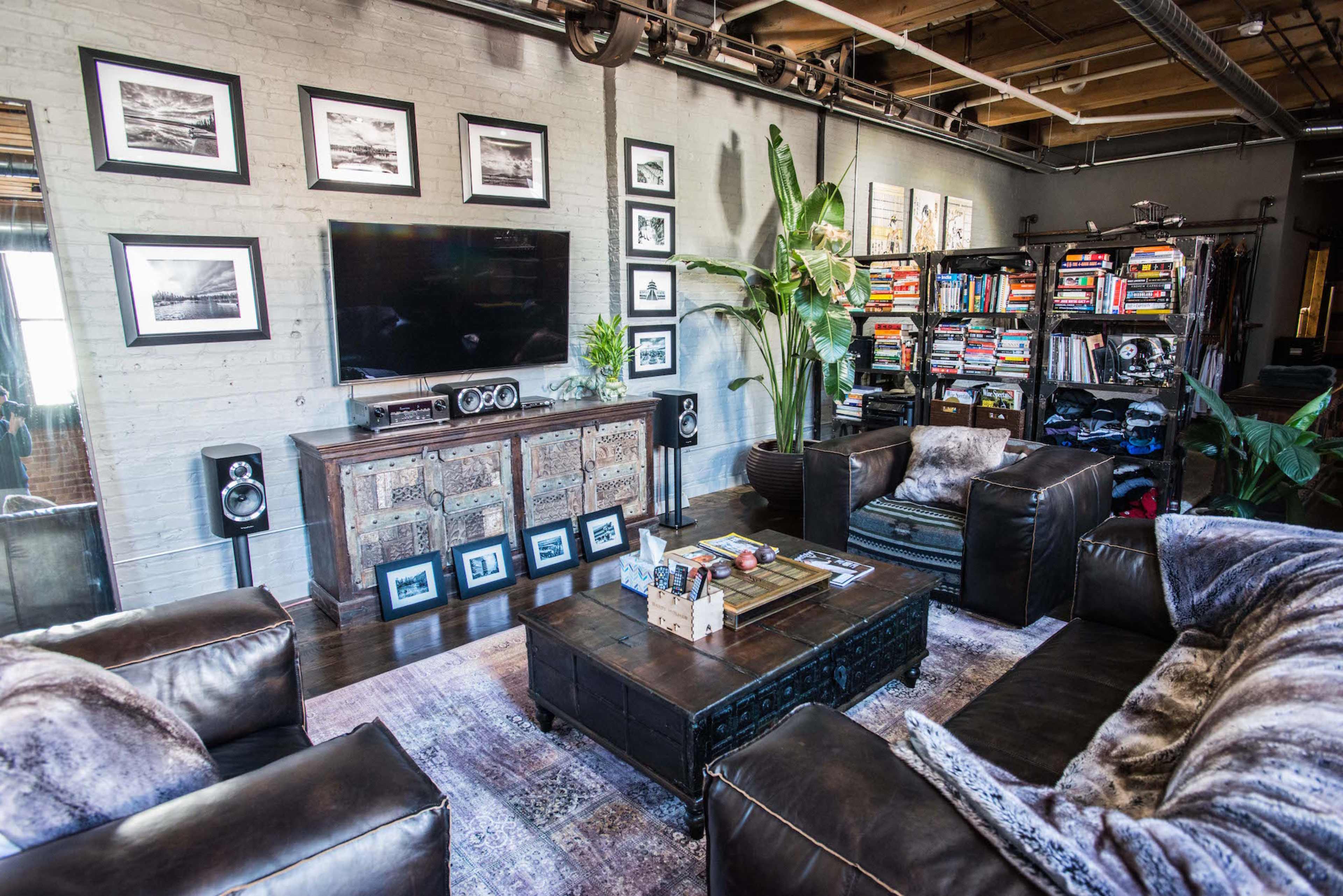 A well-furnished living room featuring a television, framed pictures on the walls, a coffee table, two armchairs, and a bookshelf filled with various books.