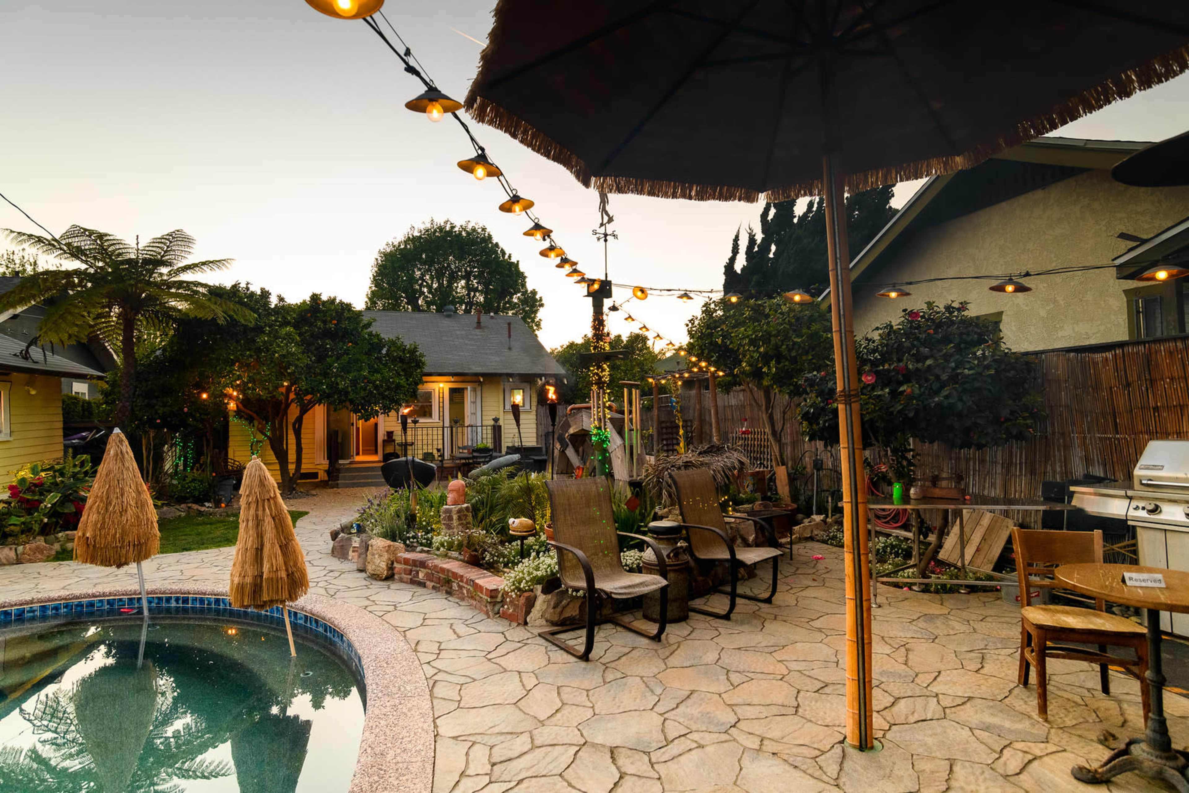 A tranquil backyard featuring a stone patio, a swimming pool, and string lights overhead, surrounded by plants and outdoor furniture.