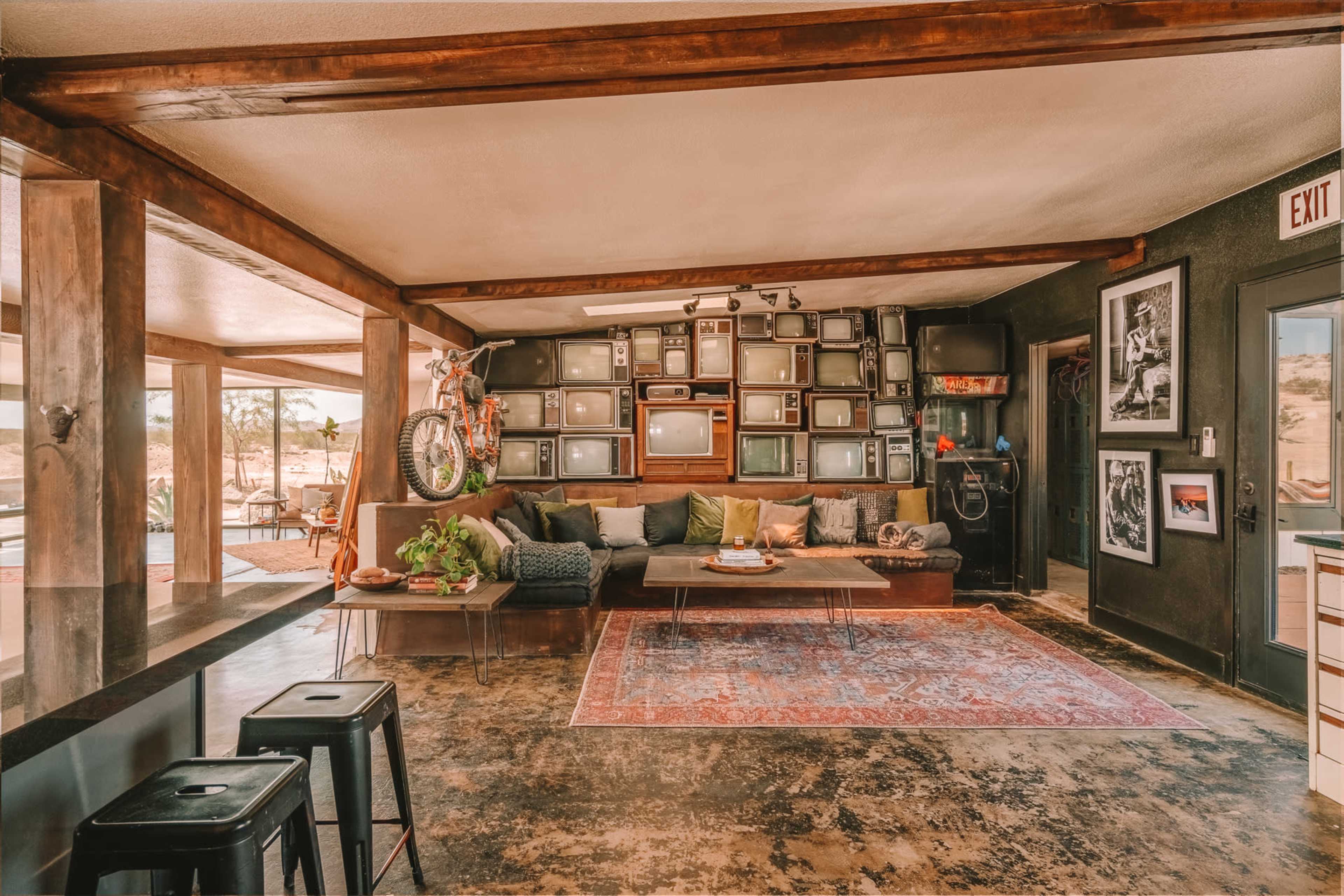 The image shows a living room with a large sectional sofa, a wall adorned with multiple vintage televisions, and a rug on a concrete floor, illuminated by natural light from large windows.