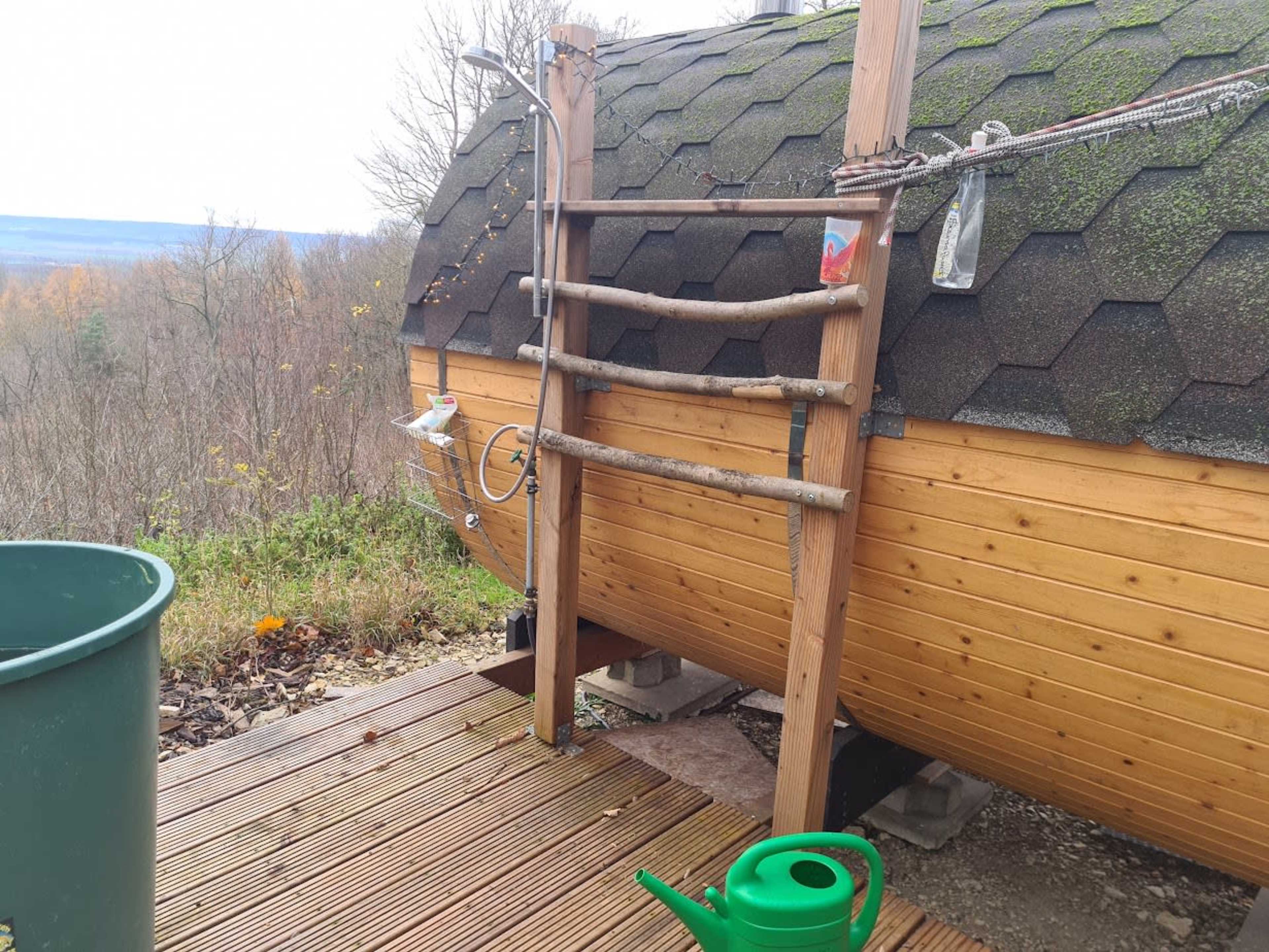 The image shows a wooden sauna with a shingled roof situated on a deck near a wooded area.