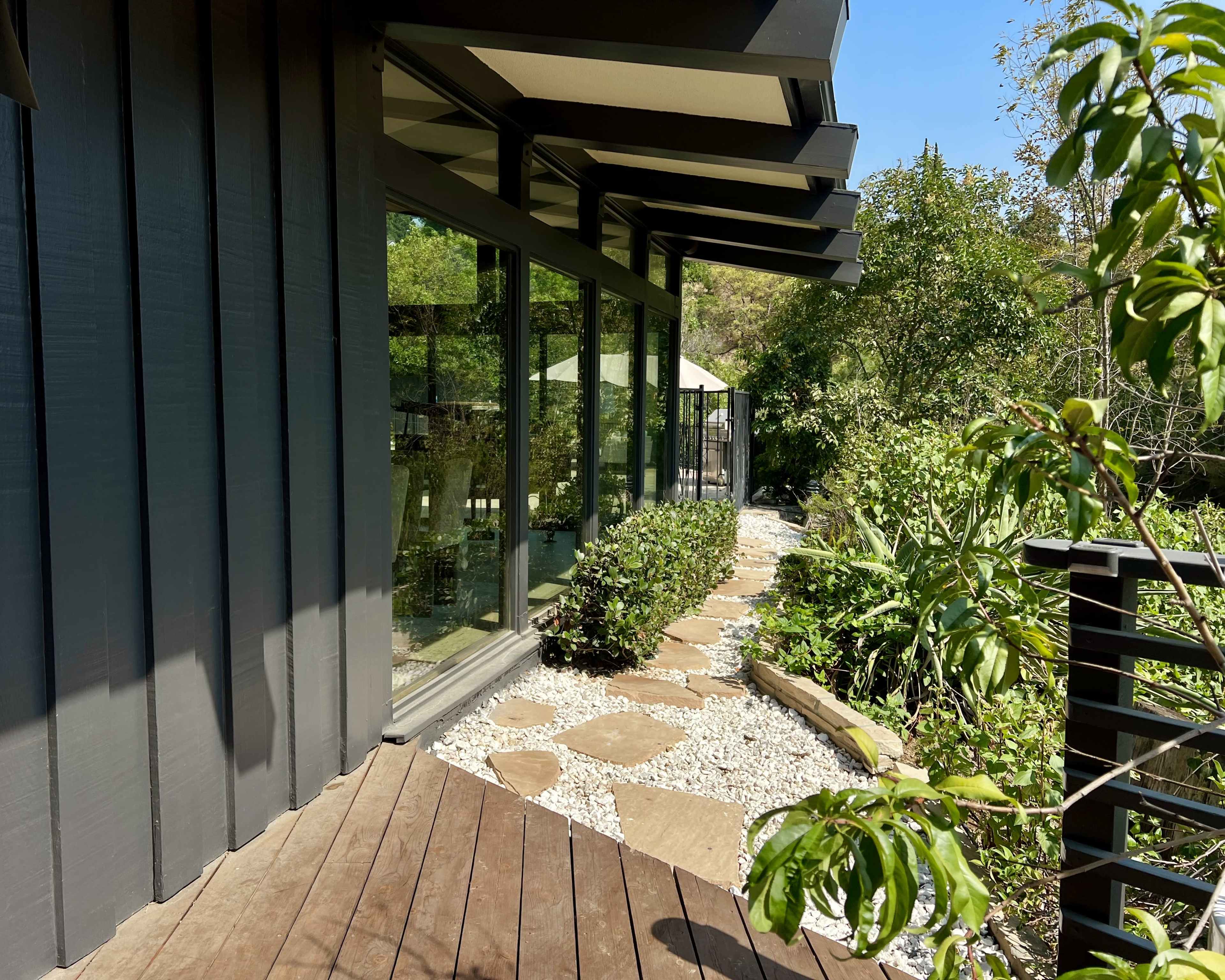 A stone pathway leads alongside a modern building with large glass windows, surrounded by lush greenery.