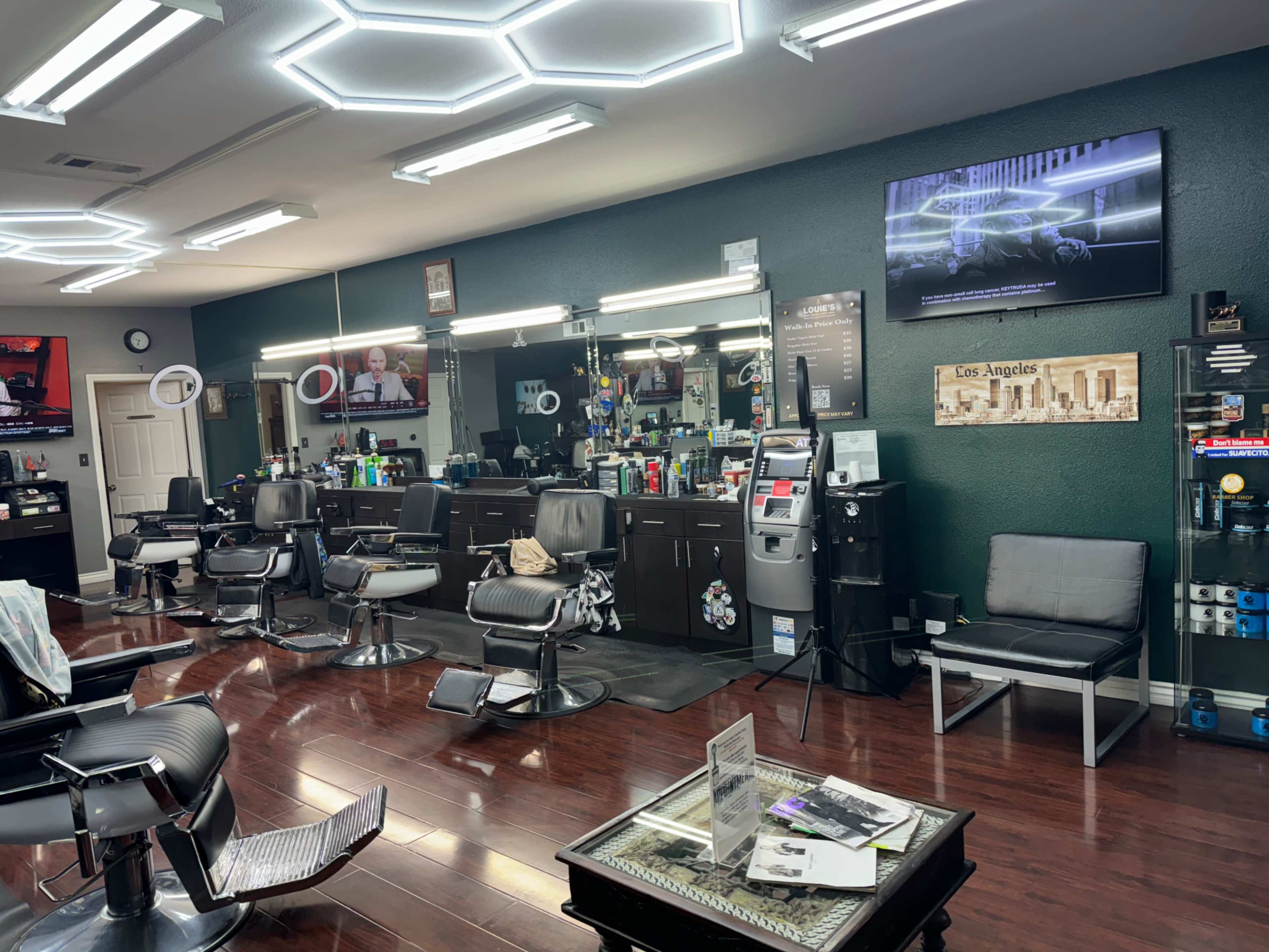 The image shows a modern barbershop interior with sleek black barber chairs, a waiting area with a gray couch, and multiple mirrors reflecting various hair grooming tools, along with a television mounted on the wall.