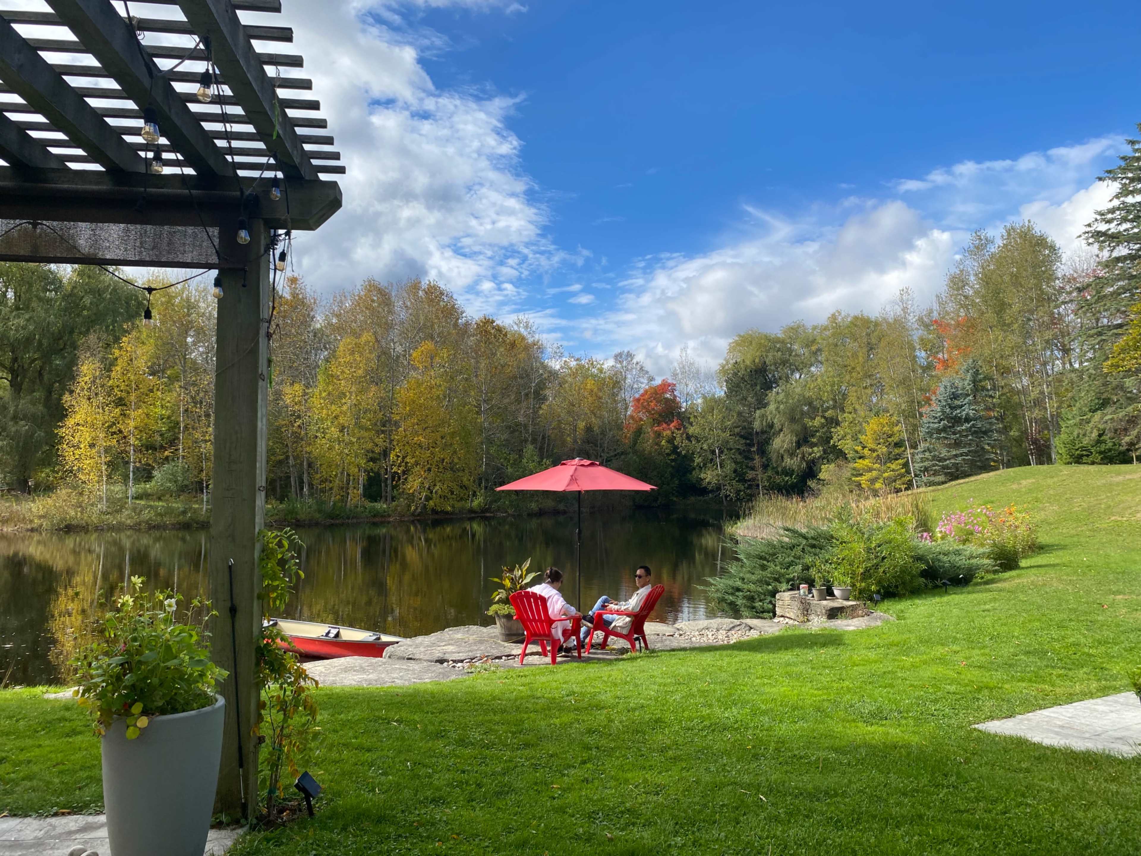 Two people relax in red chairs under a large umbrella by a pond surrounded by colorful autumn trees.