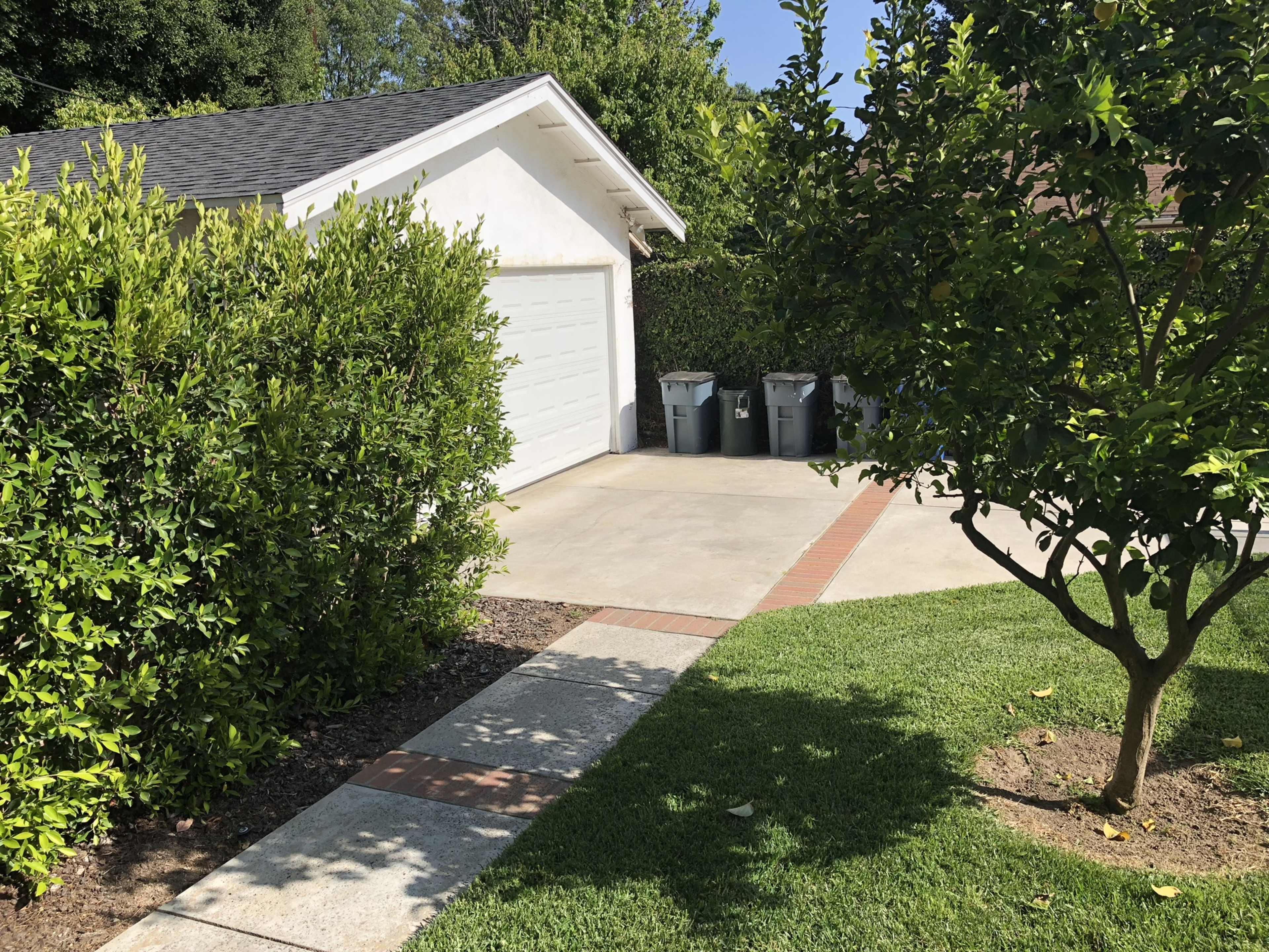 A driveway leads to a garage, with three trash bins positioned beside well-maintained hedges and a small tree.