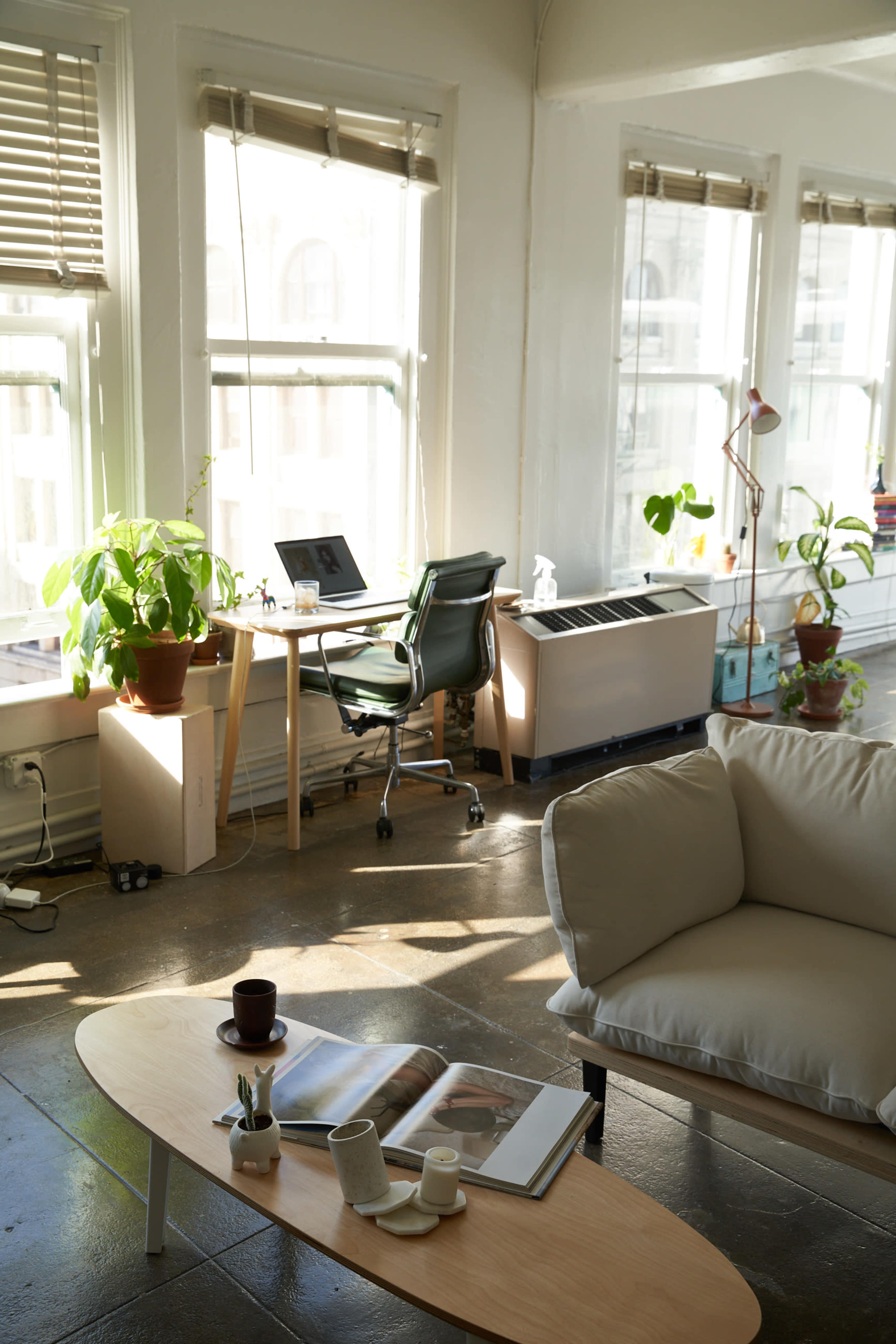 A bright, minimalist living space with a cozy sofa, a coffee table displaying books and a mug, and a desk with a computer next to large windows.