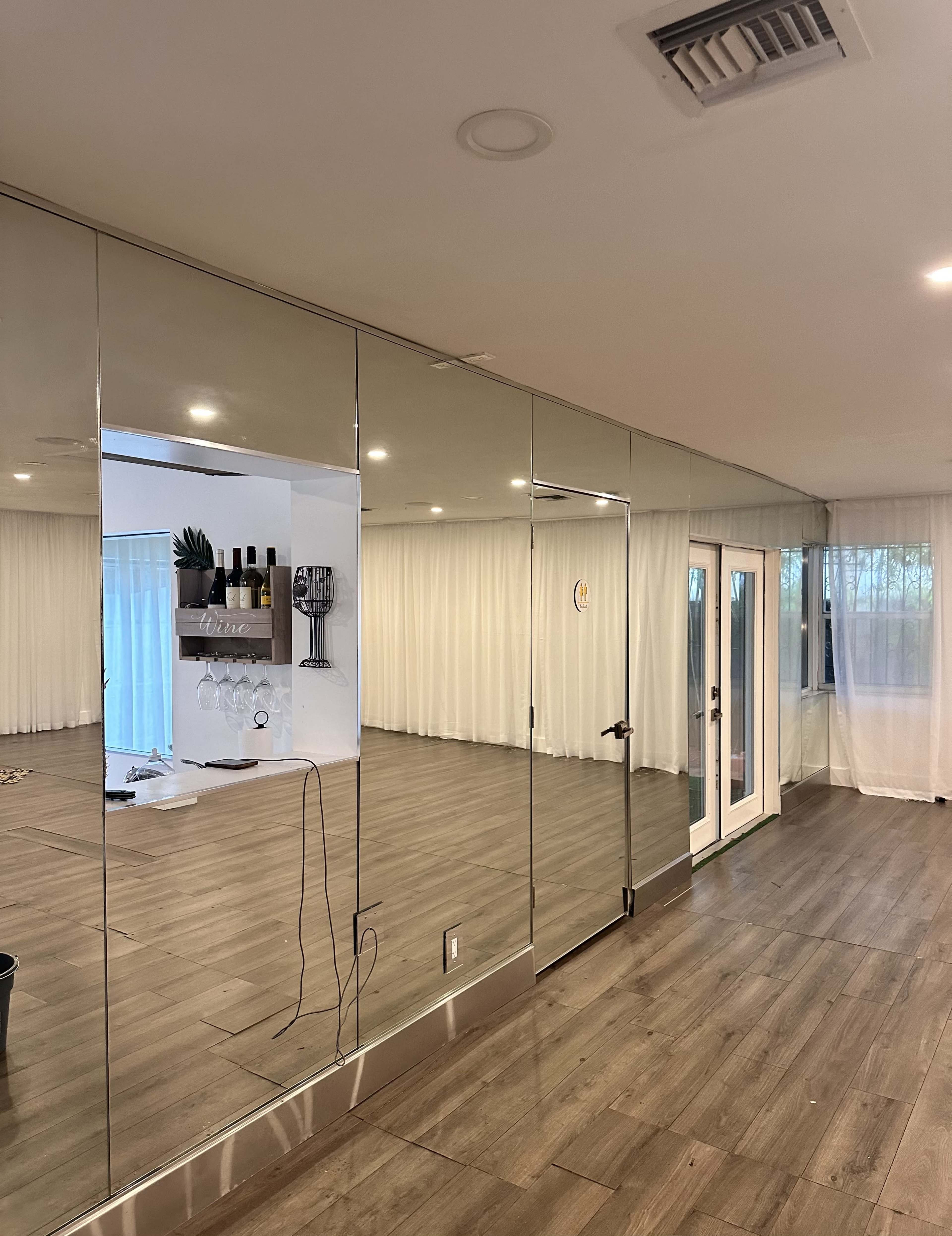 A spacious room with mirrored walls, hardwood flooring, and large glass doors leading to an outdoor area, all complemented by sheer white curtains.