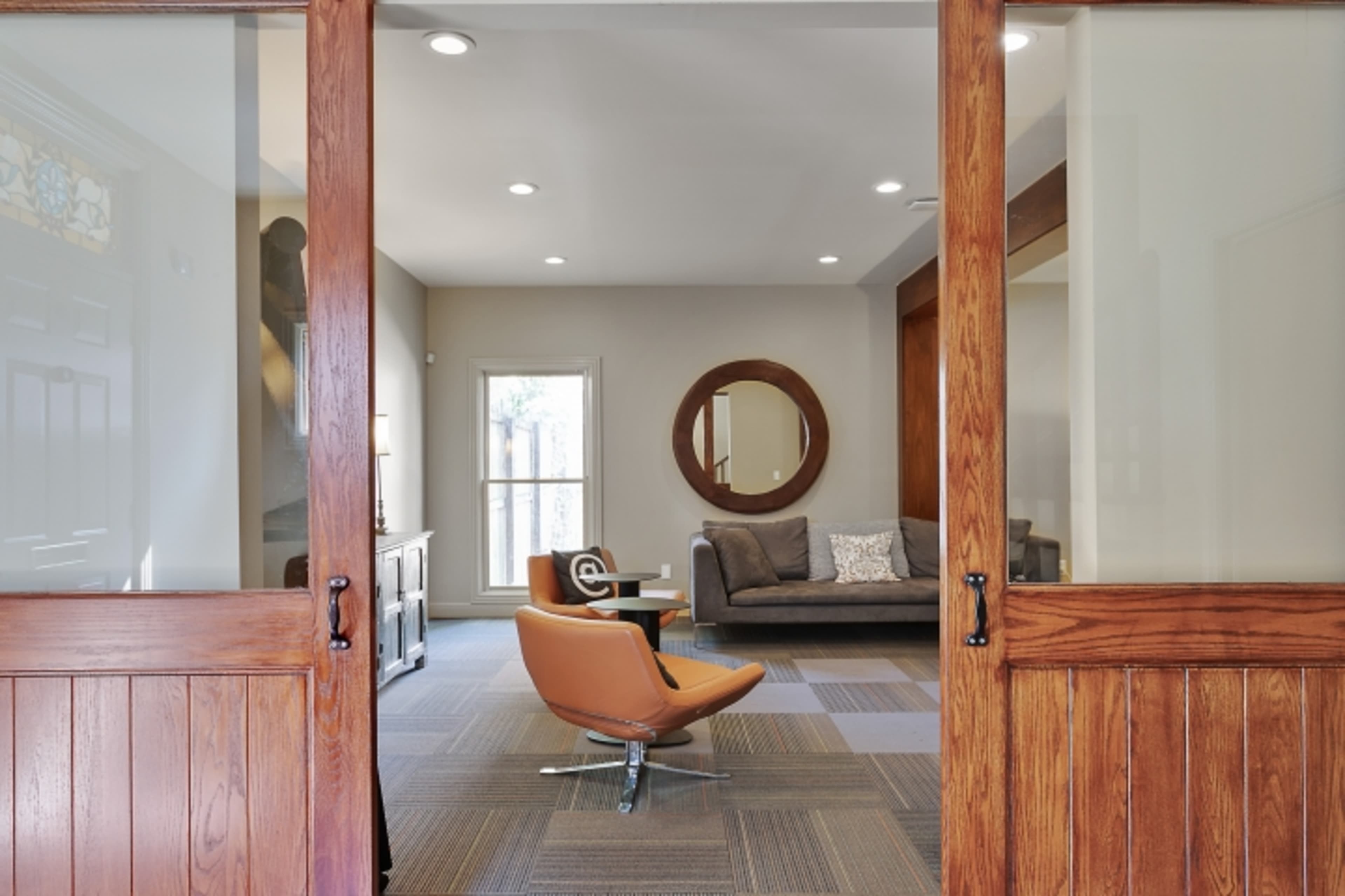 A modern lounge area is visible through glass-paneled wooden doors, featuring a gray sofa, an orange swivel chair, and a round mirror on the wall.