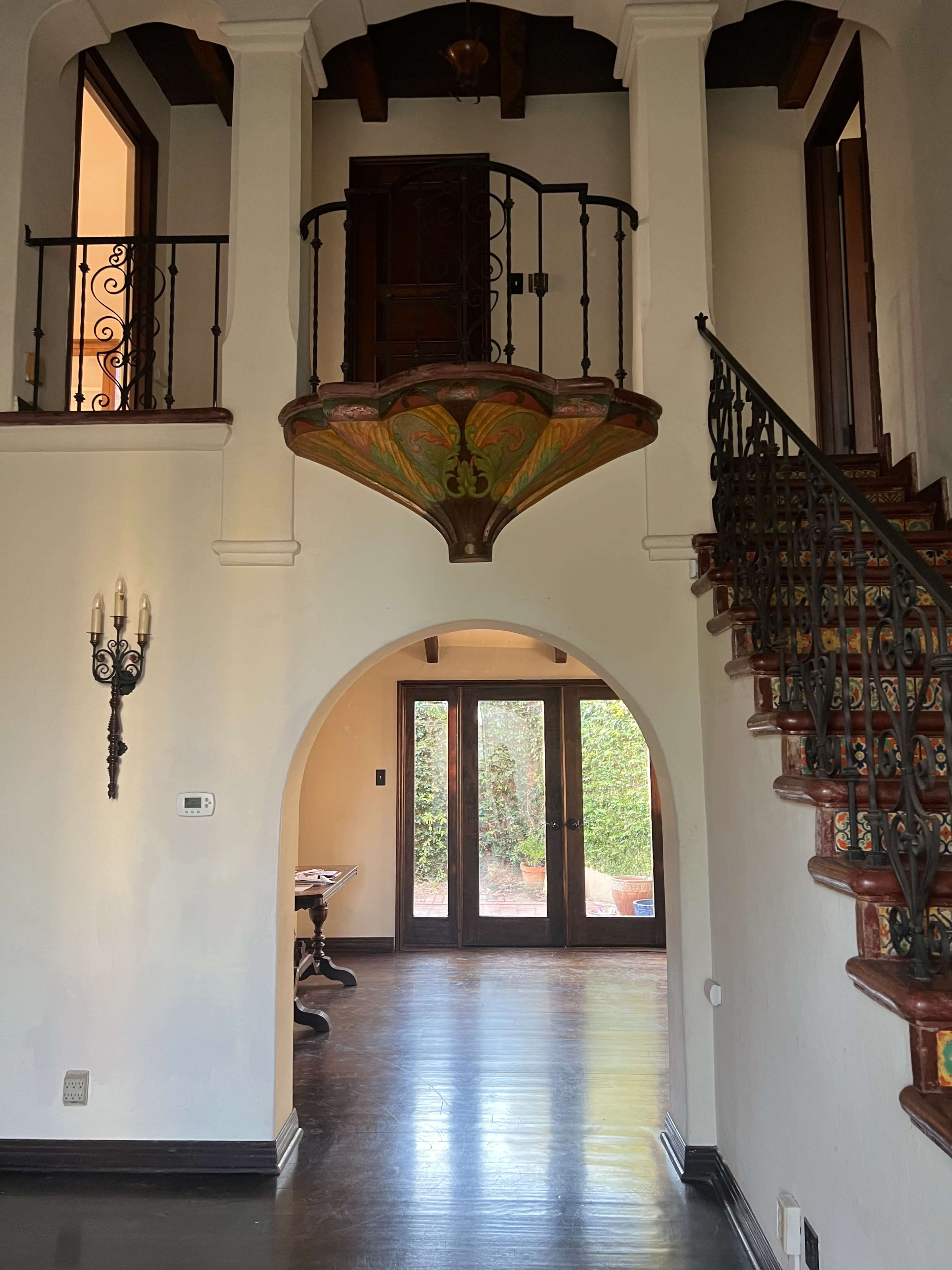The image shows an interior of a house featuring a staircase with ornate iron railings, a second-floor balcony with decorative elements, and large windows leading to an outdoor area.