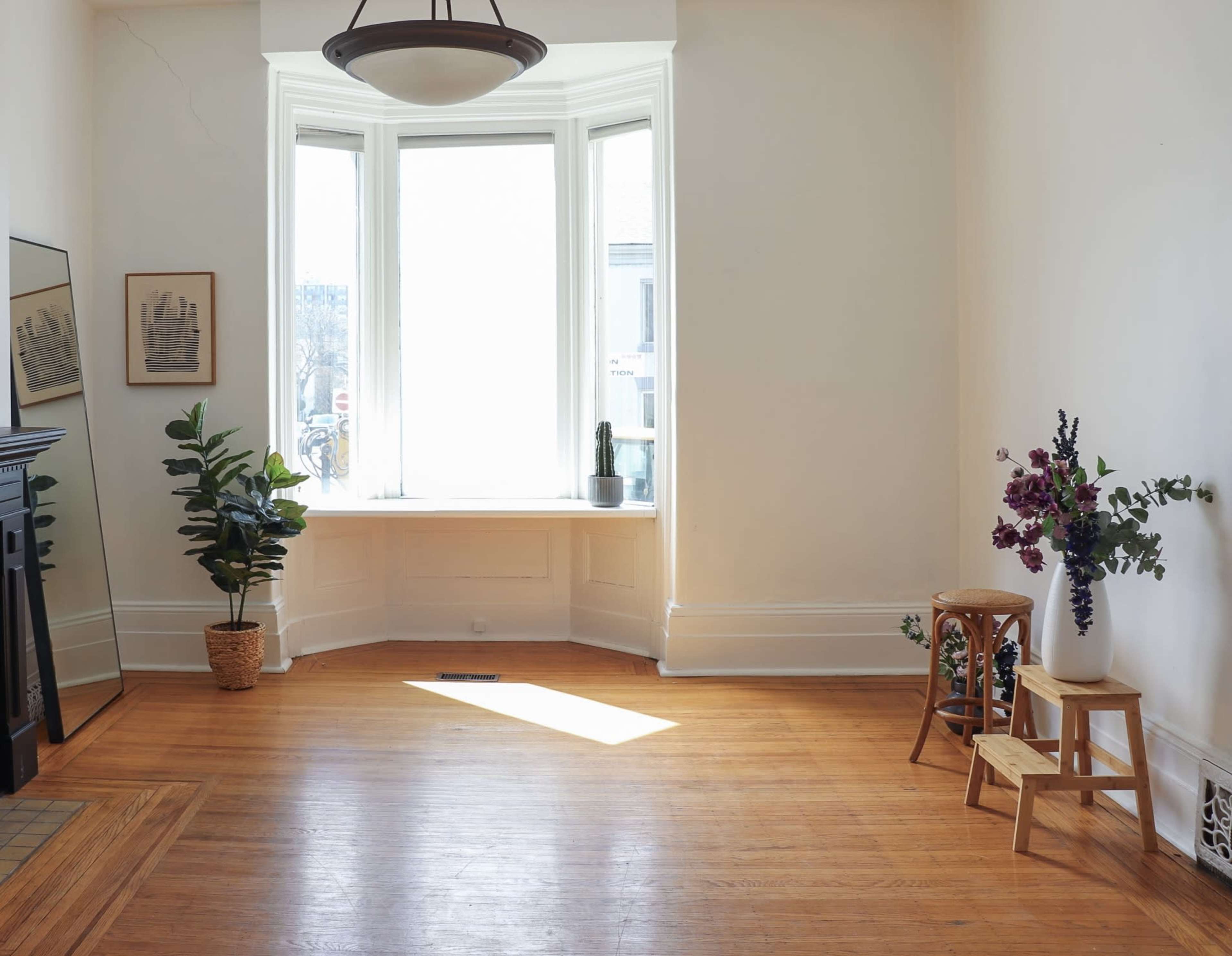 The image shows a bright and airy room with a bay window, hardwood floors, a tall plant in a pot, a small wooden stool, and a bouquet of flowers in a corner.