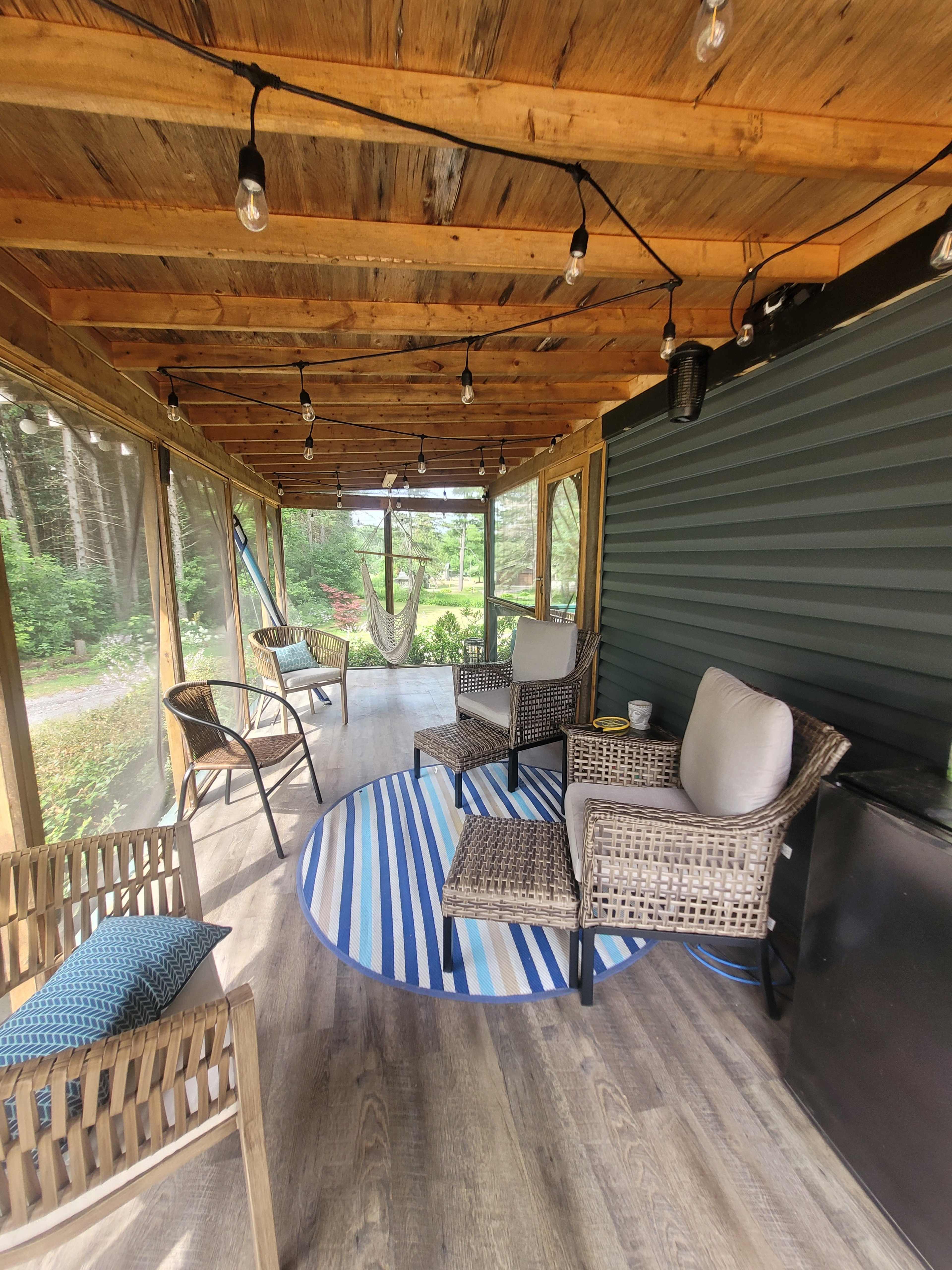 The image shows a screened-in porch with wooden beams, woven chairs, a round blue and white rug, and string lights, surrounded by greenery.