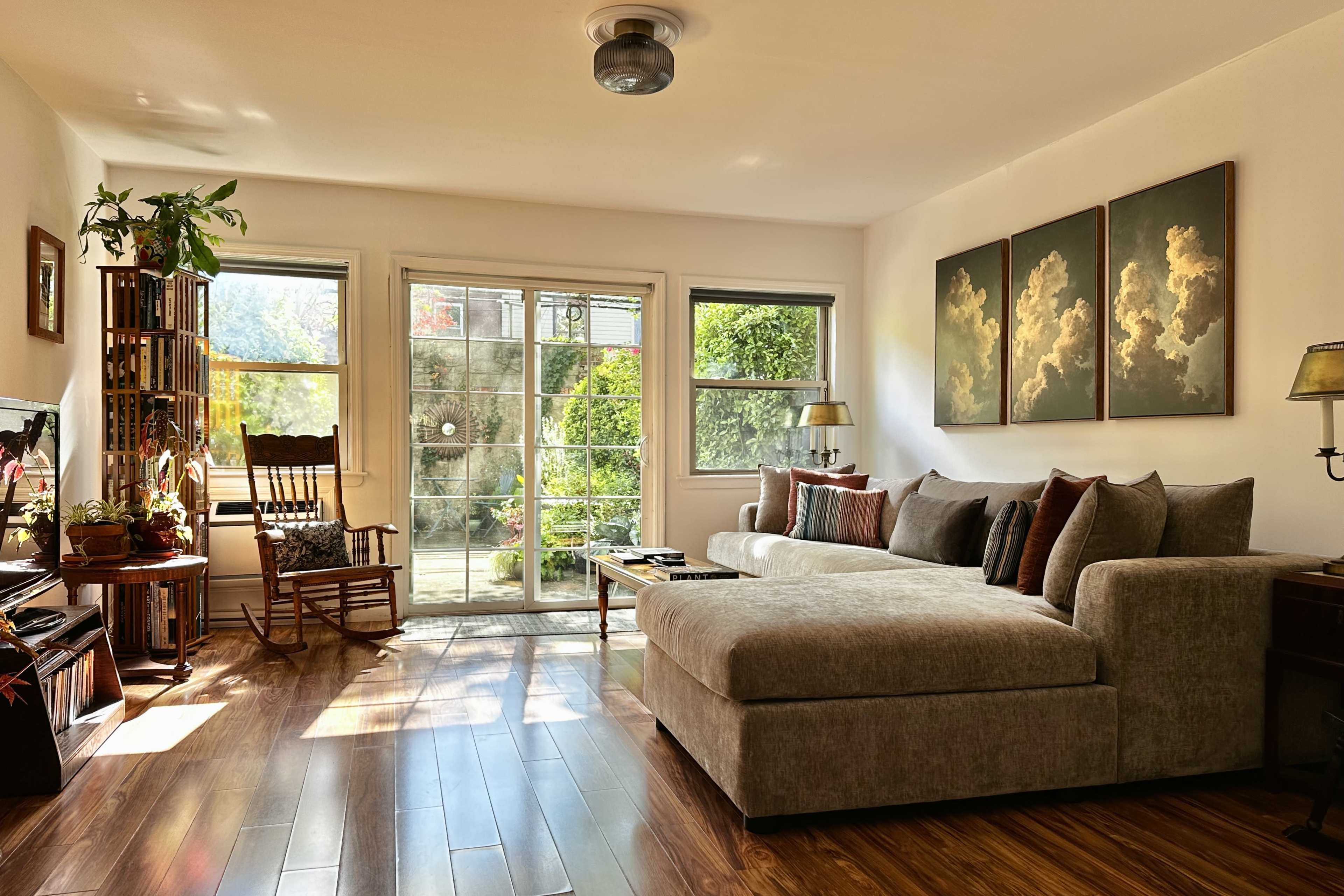 The image shows a cozy living room with a large sectional sofa, a rocking chair, and a view of a garden through glass sliding doors.