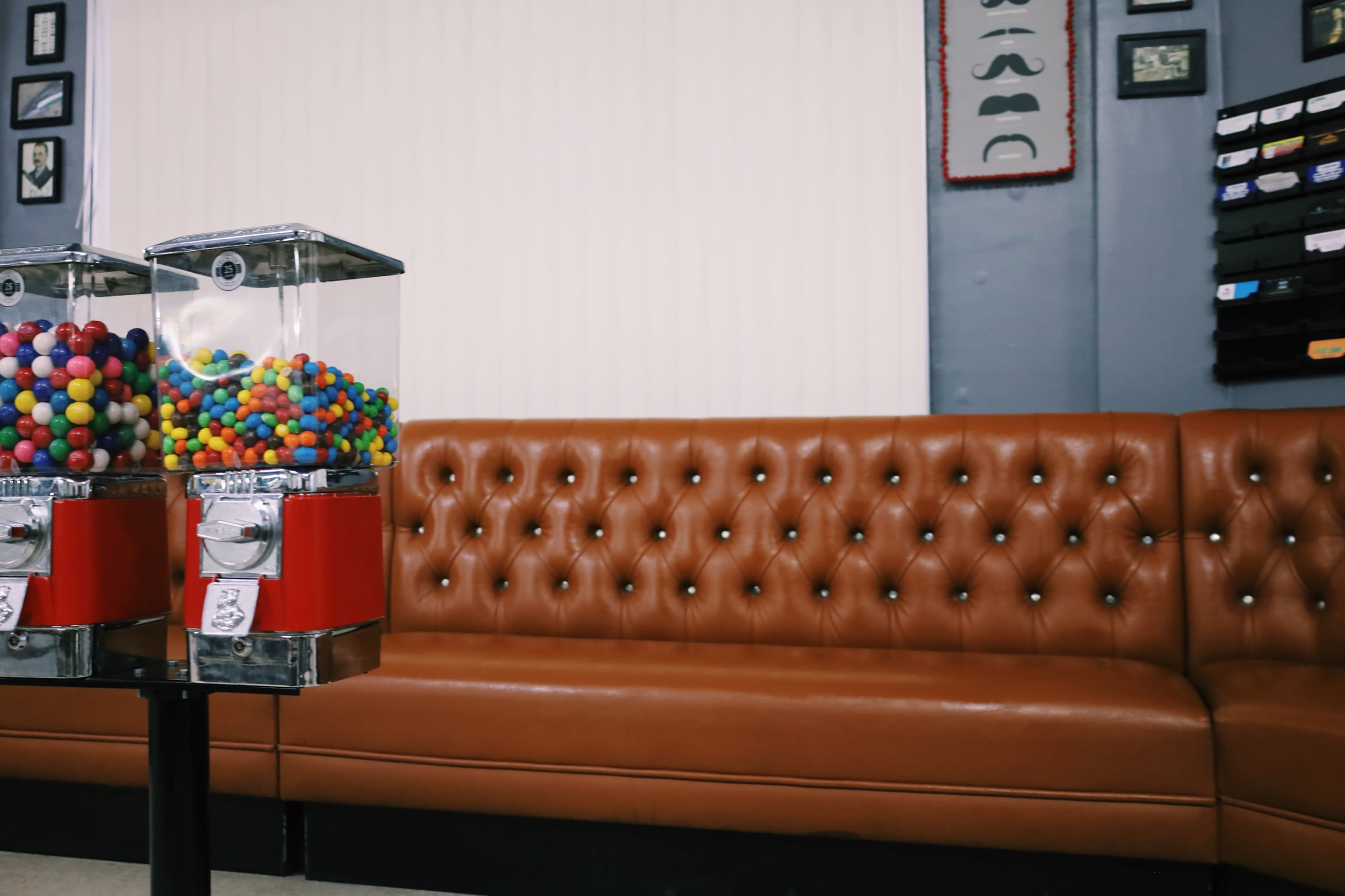 The image shows two gumball machines beside a brown tufted sofa in a room with plain walls and various decorations.