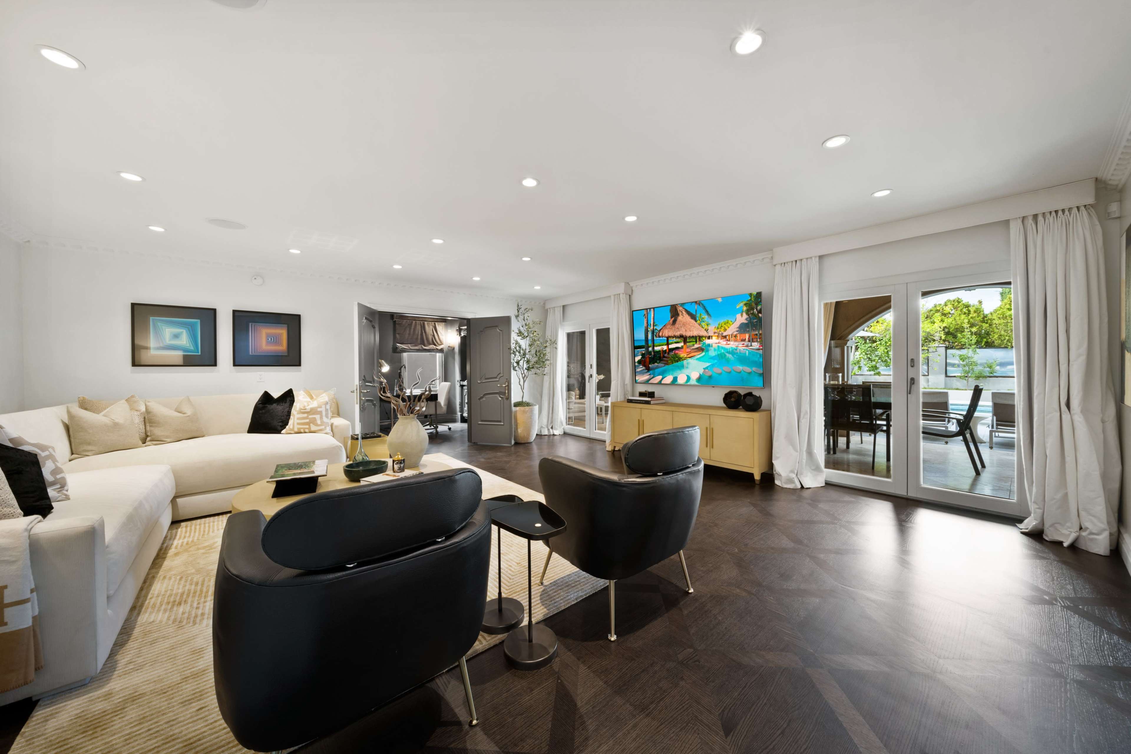 The image shows a modern living room with a large sectional sofa, two black chairs, and a wall-mounted TV displaying a poolside scene, with glass doors leading to an outdoor area.