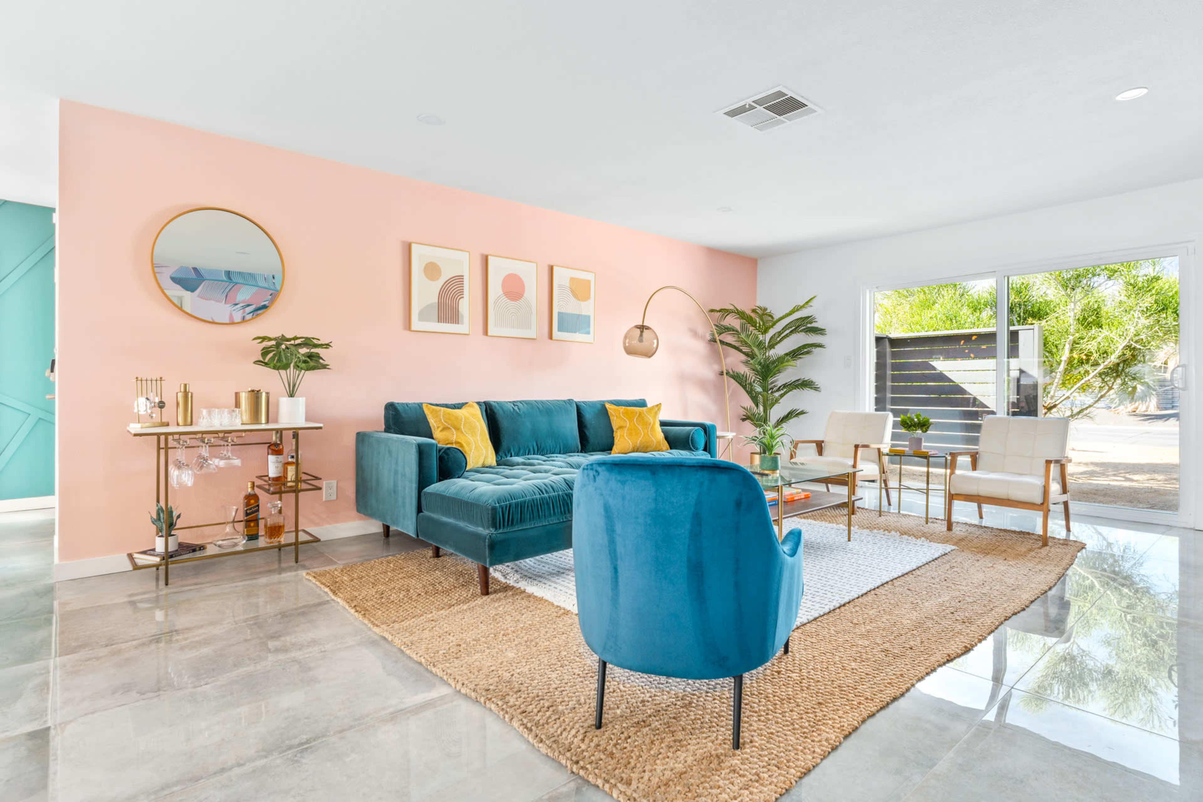 A bright living room features a teal velvet couch, a round blue chair, light-colored armchairs, a jute rug, and large windows opening to a patio.