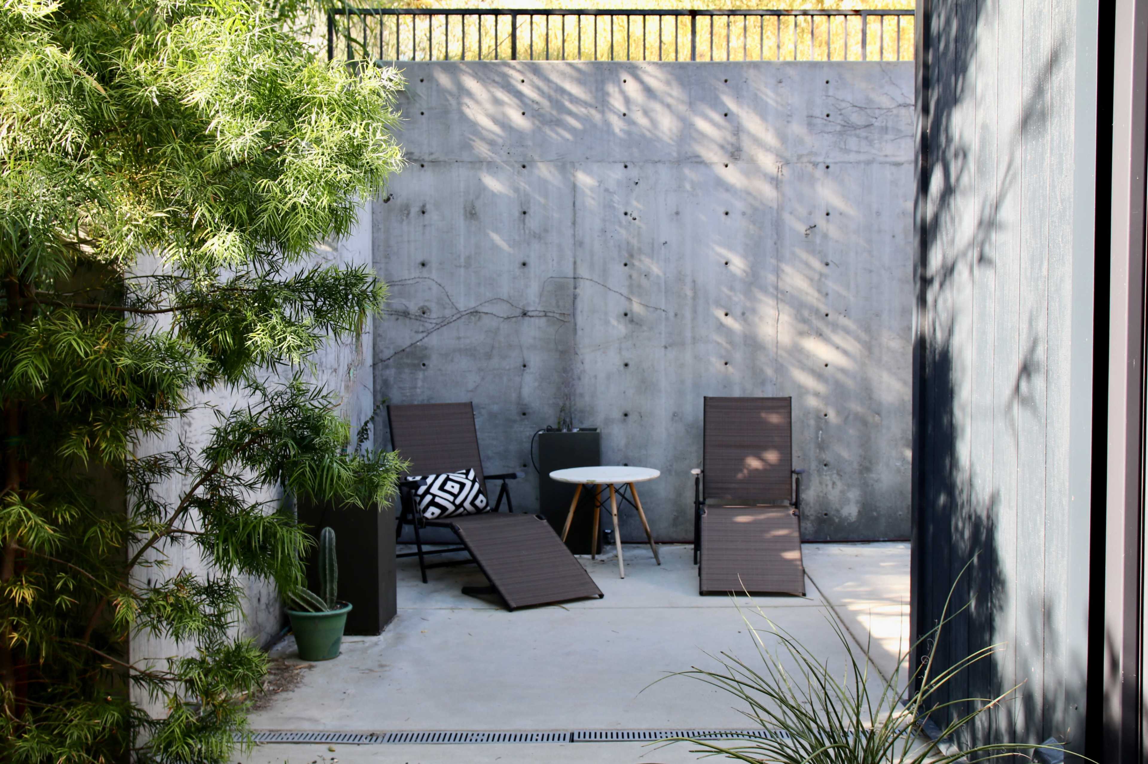 The image shows a modern outdoor space featuring two lounge chairs, a small round table, and a potted plant, surrounded by a concrete wall and greenery.