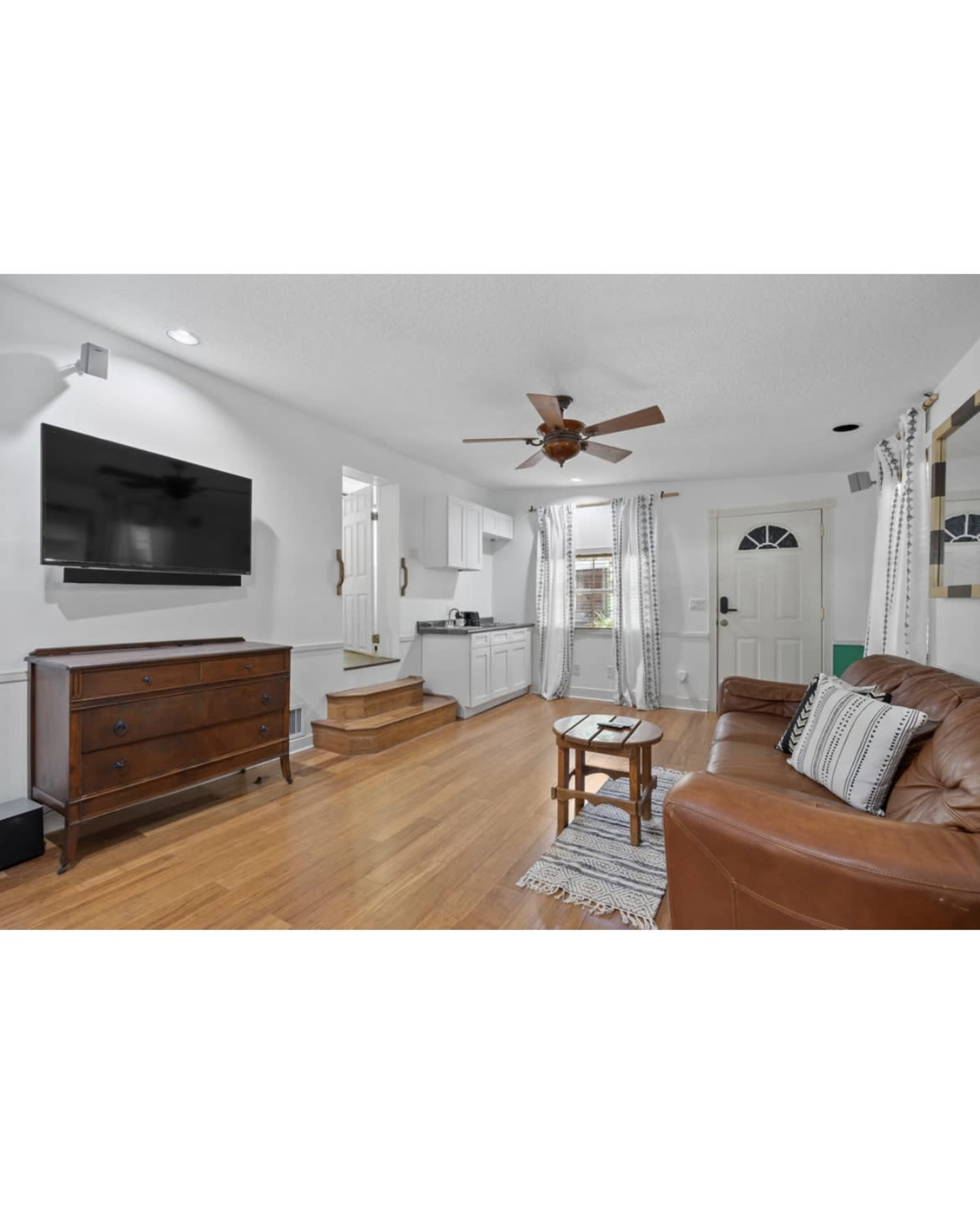 The image shows a living room featuring a leather couch, wooden furniture, a wall-mounted TV, and a staircase leading to another area.