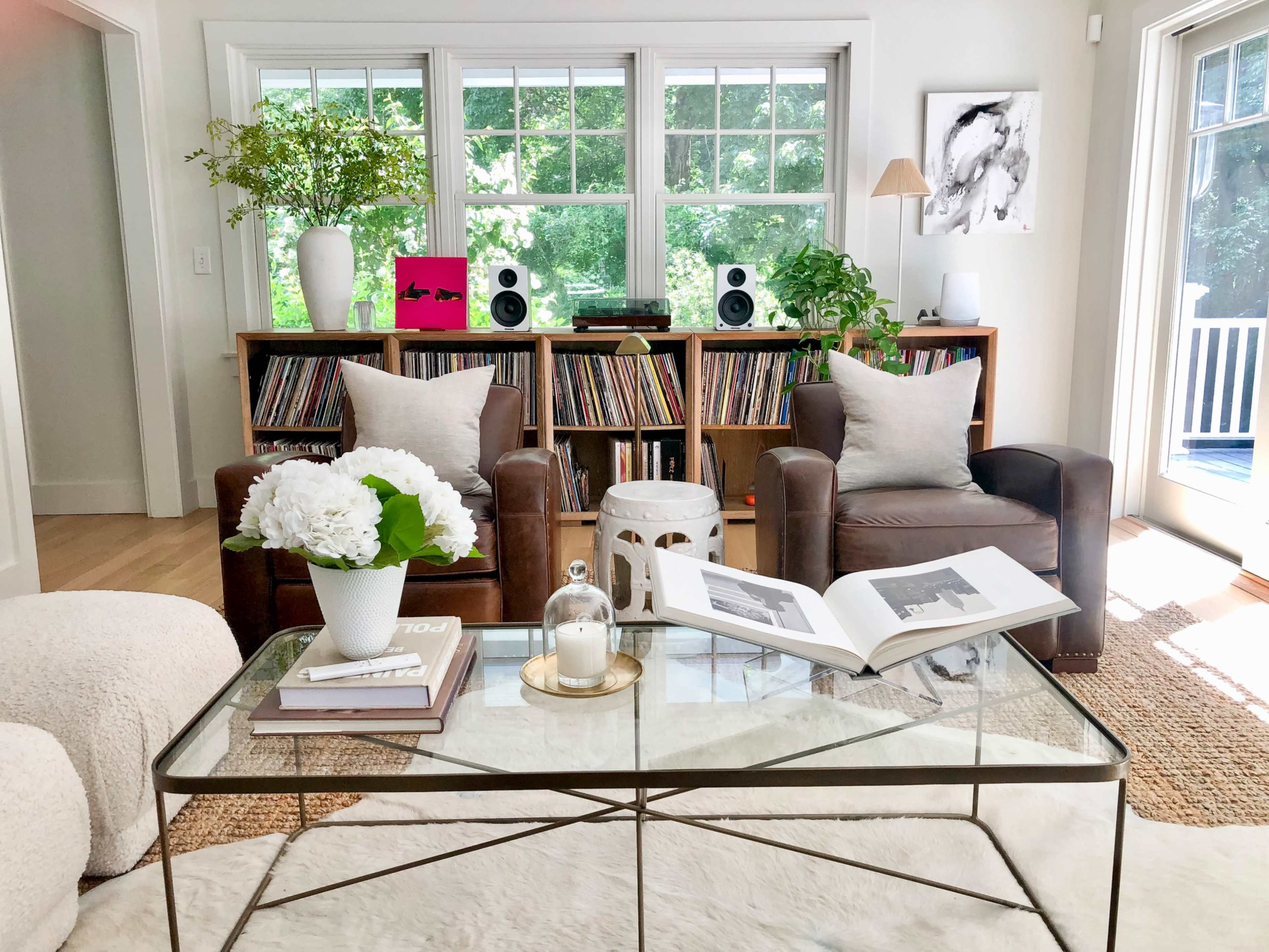 The scene features a cozy living room with two leather armchairs, a glass coffee table, and a wooden bookshelf filled with records, all illuminated by natural light from large windows.
