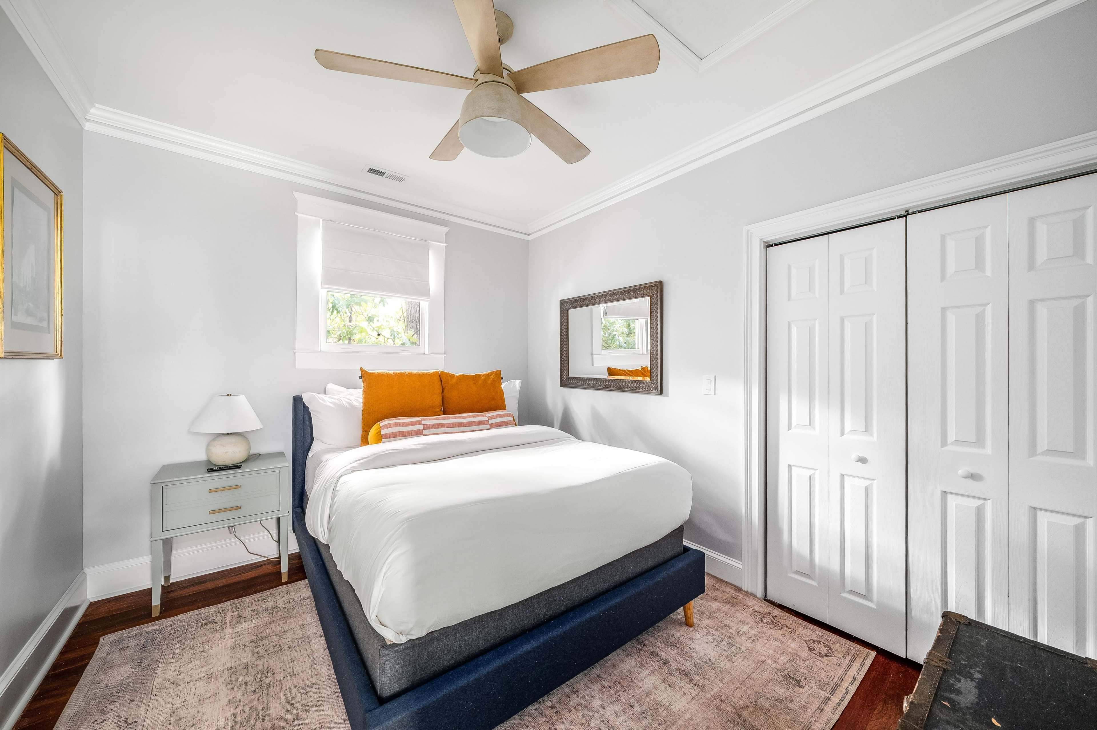 The image shows a neatly arranged bedroom with a bed adorned with white bedding and orange accent pillows, a nightstand with a lamp, and double white closet doors.