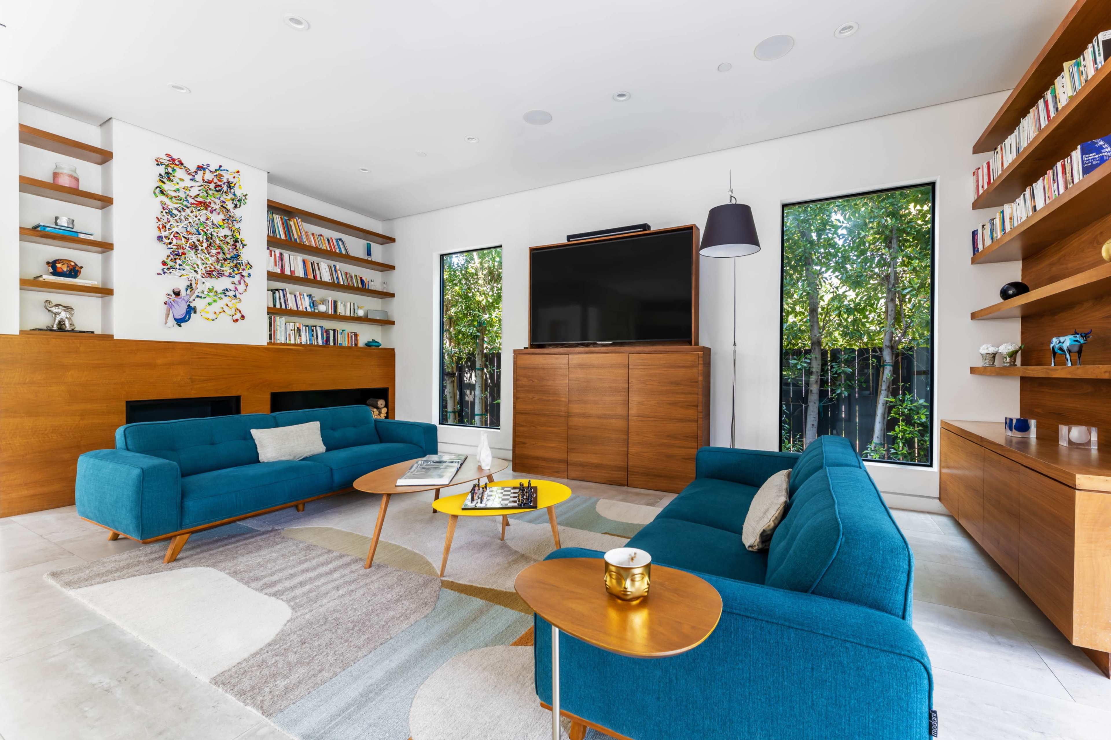 The room features modern teal sofas, a coffee table with a yellow accent, and large windows that offer a view of greenery, with shelves filled with books.